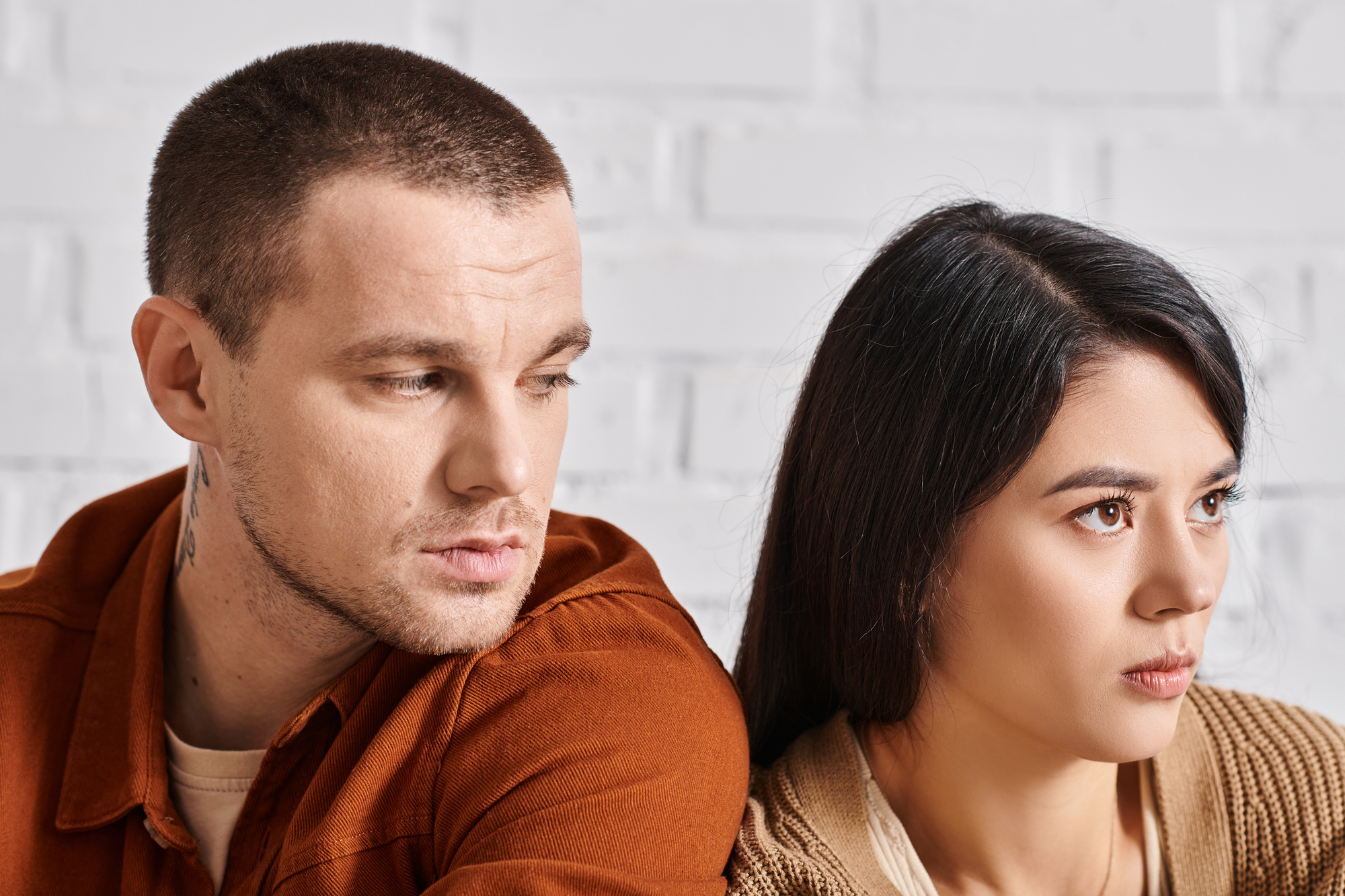 A close-up of a man and a woman sitting together against a white brick wall, both looking in different directions. The man, in an orange shirt, appears contemplative, while the woman, in a tan sweater, has a serious expression.