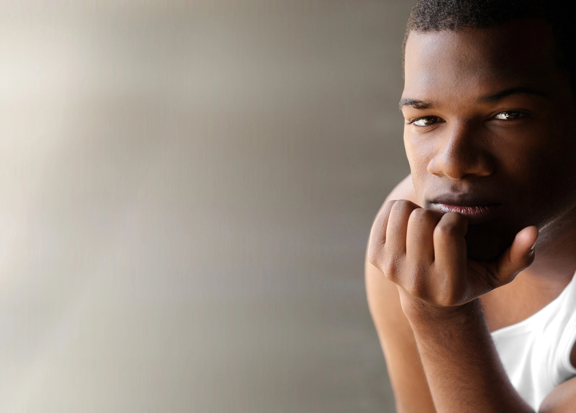 A young person with short hair, wearing a white sleeveless shirt, rests their chin on their hand and looks thoughtfully at the camera. The background is blurred and neutral, drawing attention to the subject's contemplative expression.