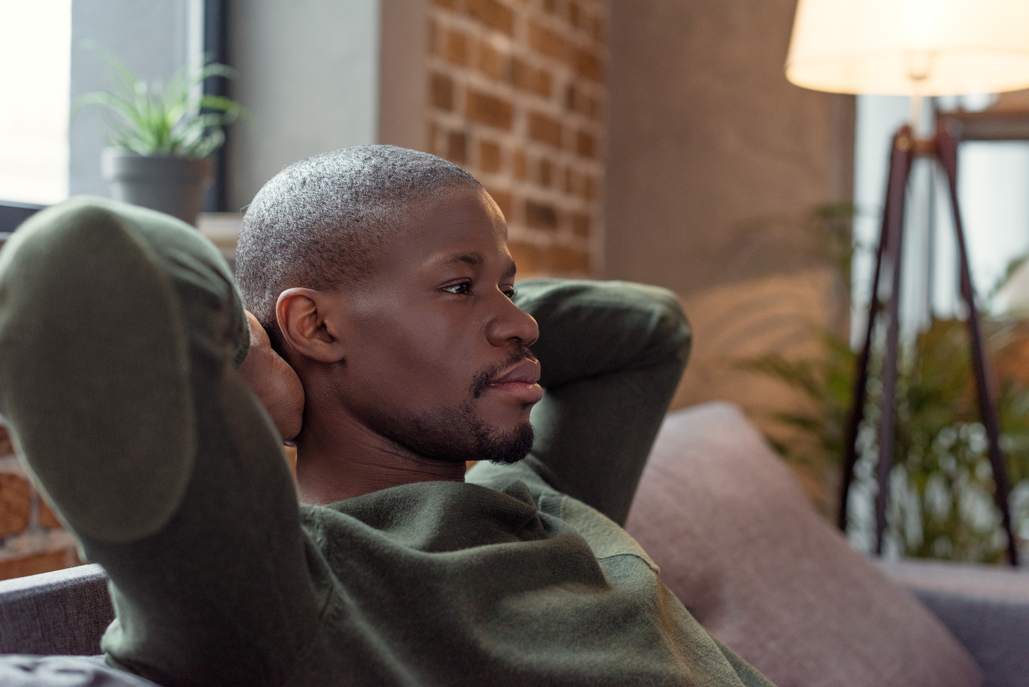 A young man with short hair and a green sweater is sitting on a couch with his hands resting behind his head. He is looking calmly to the side in a cozy living room with brick walls, indoor plants, a window, and a standing lamp in the background.