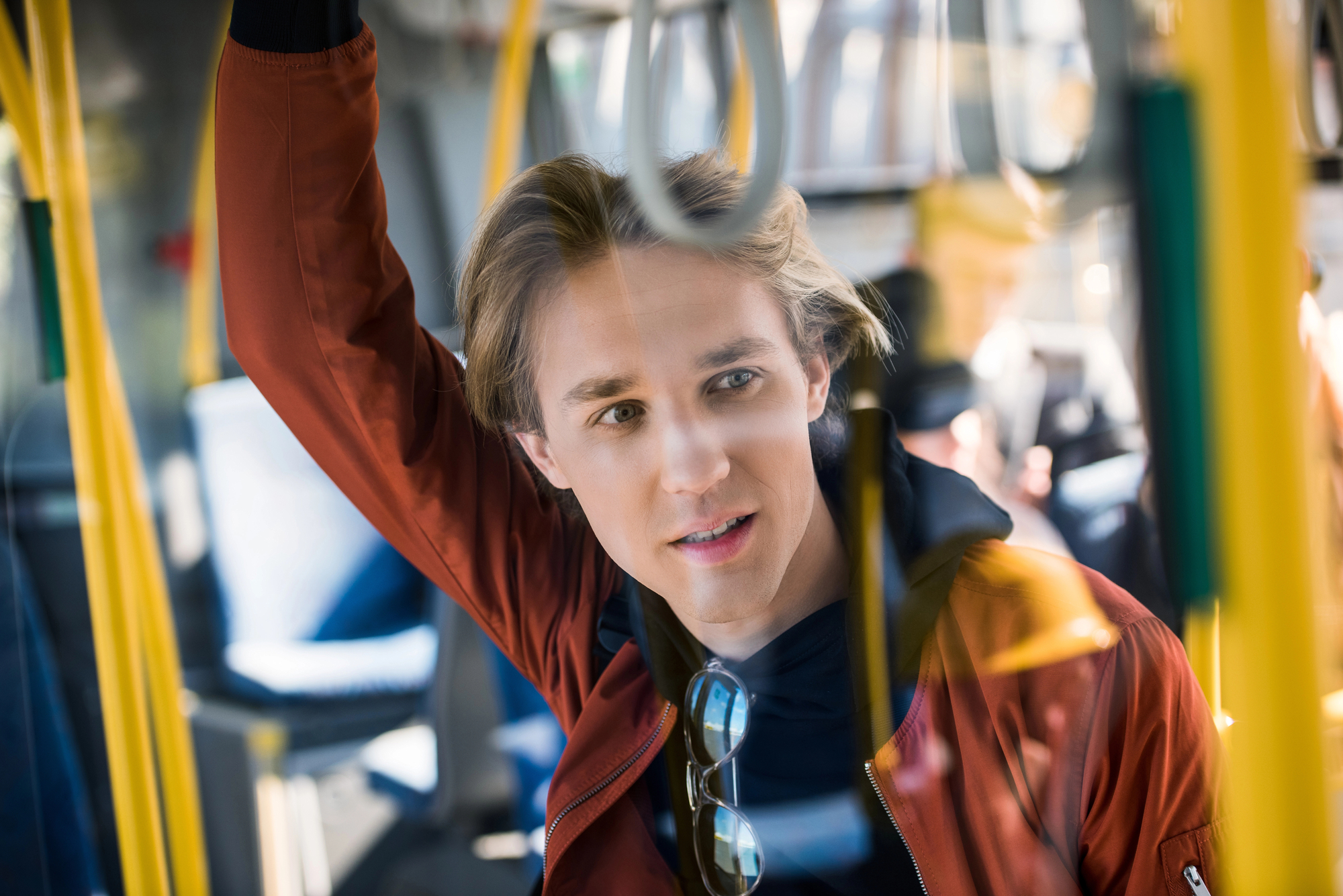 A young man with light brown hair, wearing a red jacket and a pair of sunglasses hanging from his shirt, is standing inside a bus, holding an overhead handrail. He is looking ahead with a thoughtful expression. The bus interior and other passengers are visible.