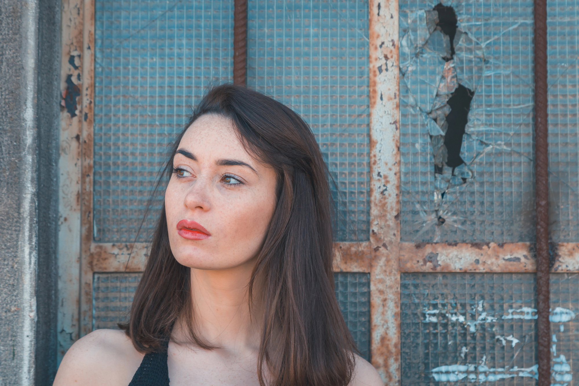 A woman with long brown hair and red lipstick is standing in front of a weathered, broken window with a metal grid. She is looking to her left, with a thoughtful expression. The background shows signs of decay and rust, creating a contrast with her appearance.