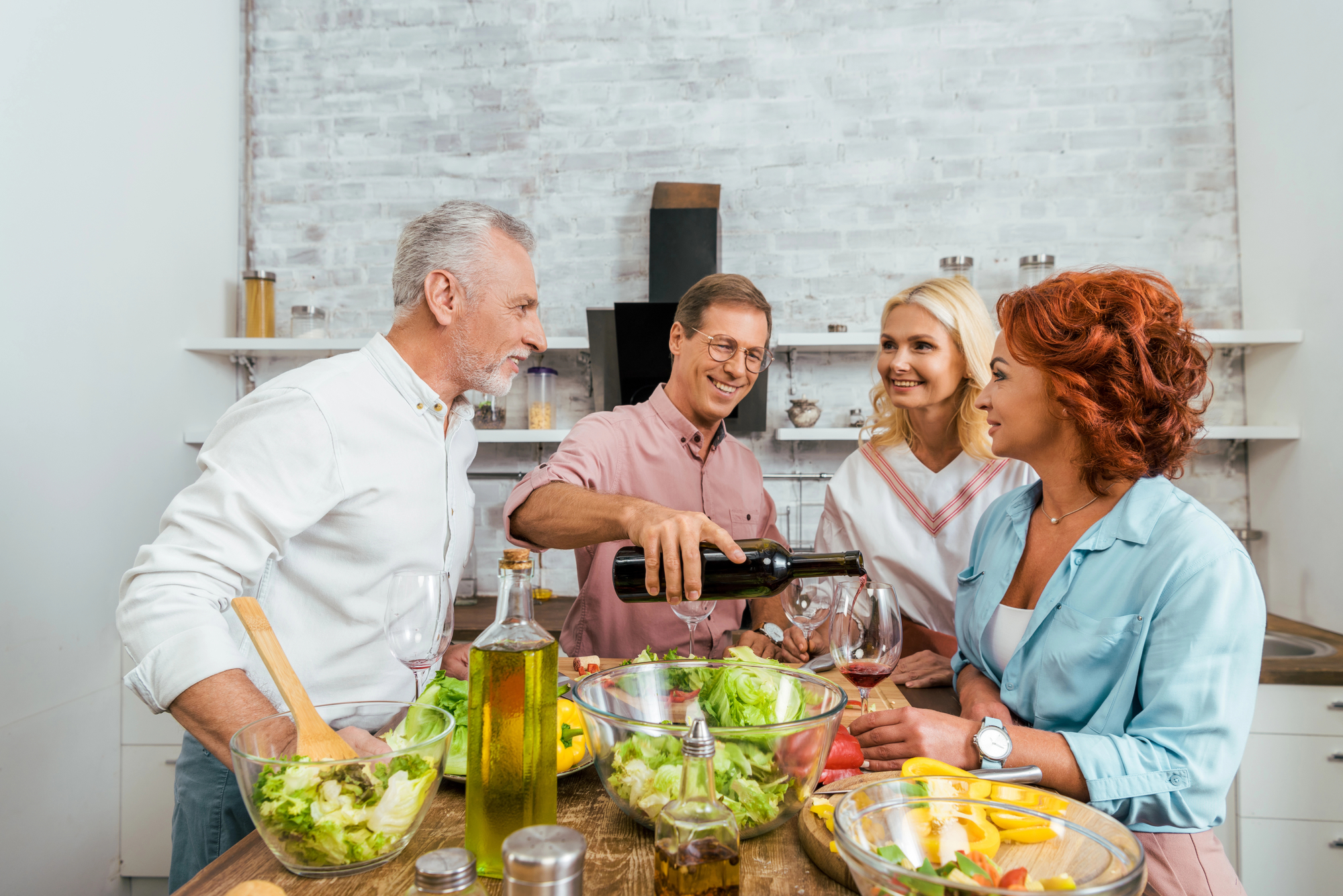 Four adults, two men and two women, stand around a kitchen island preparing a meal. One man is pouring wine into a glass. There are various bowls of salad, bottles of oil, and vegetables on the counter. The group appears to be enjoying each other's company.