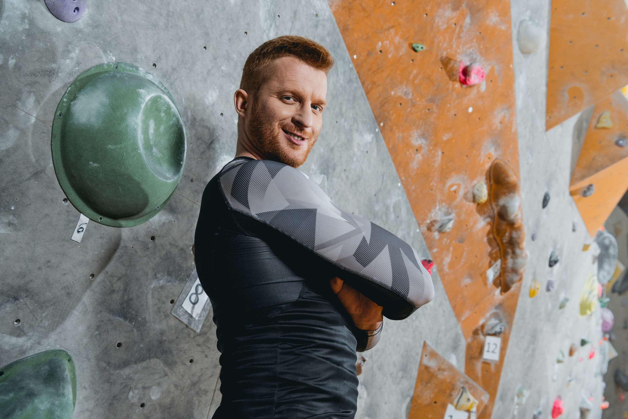 A person with red hair and a beard is standing with crossed arms in front of a rock climbing wall. They are wearing a black and grey geometric-patterned long-sleeve shirt and appear to be smiling confidently. Various climbing holds are visible on the wall.