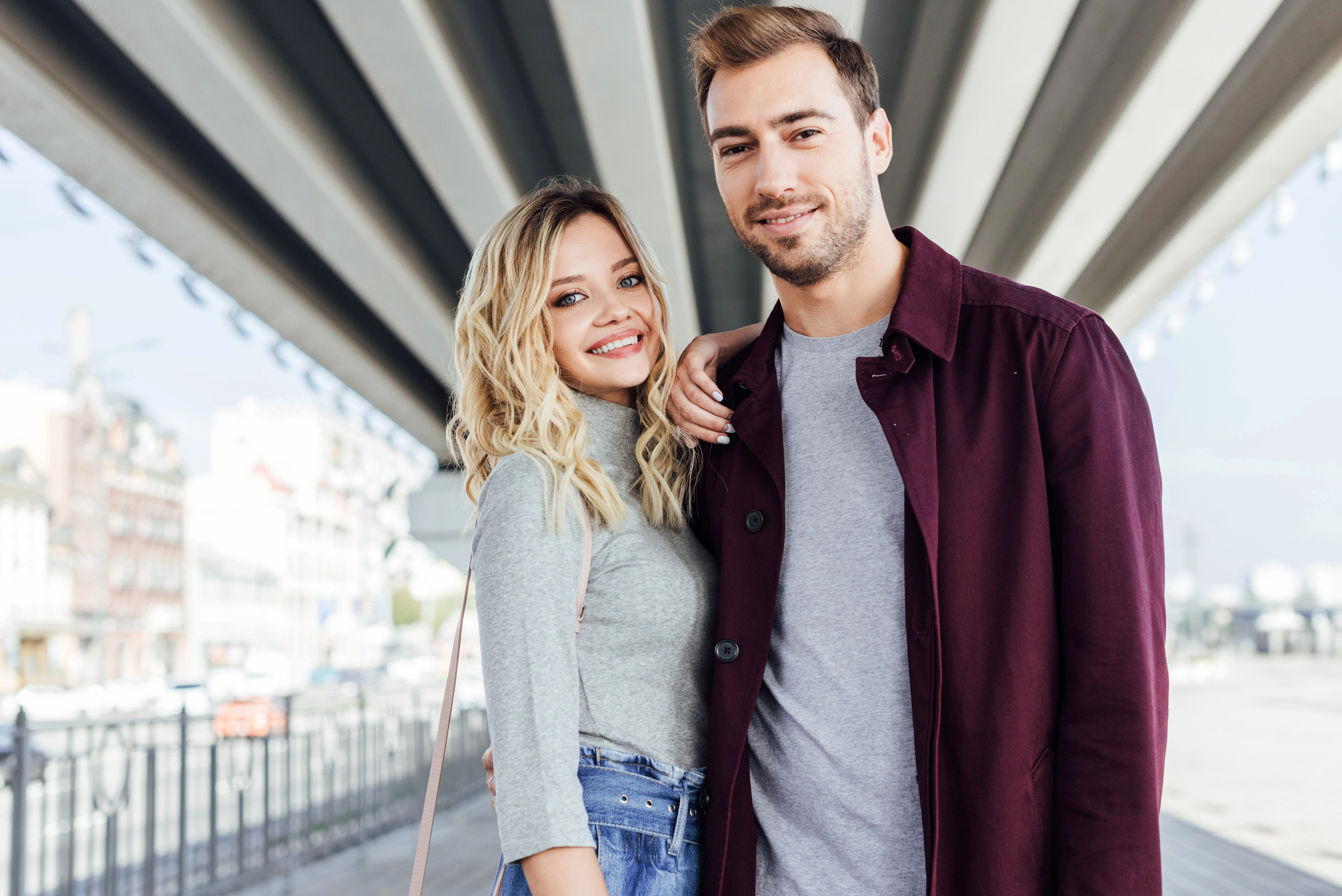 A young couple stands together under a bridge, smiling at the camera. The woman, with shoulder-length blonde hair, wears a gray top and jeans, while the man, with short brown hair, wears a gray shirt and a maroon jacket. The background features an urban setting.