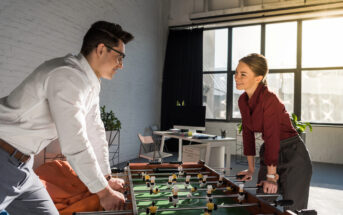 Two office workers, a man in a white shirt and a woman in a red blouse, play foosball in a brightly lit office space with large windows. The room features modern decor with a computer desk and plants in the background. They are smiling and enjoying the game.