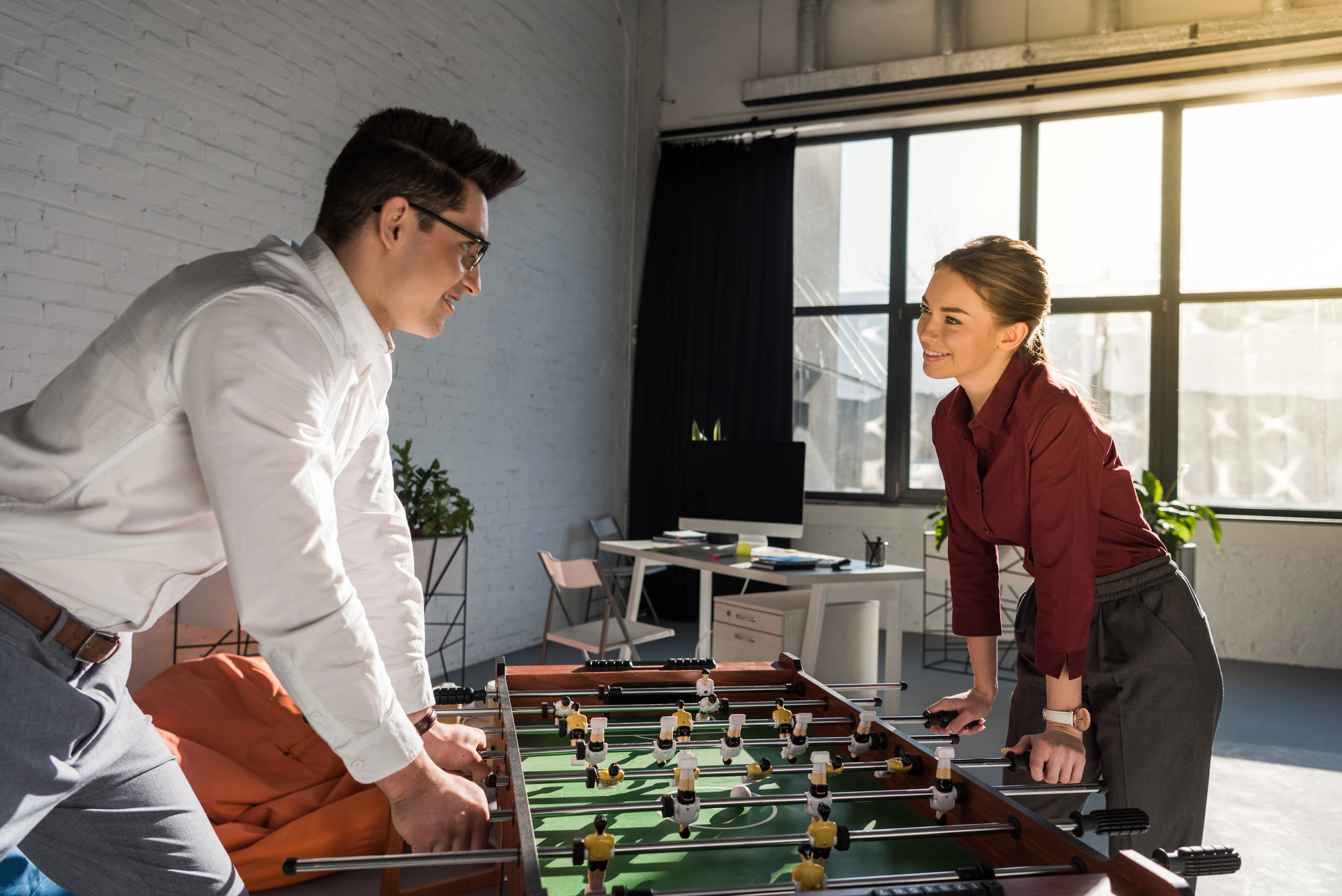 Two office workers, a man in a white shirt and a woman in a red blouse, play foosball in a brightly lit office space with large windows. The room features modern decor with a computer desk and plants in the background. They are smiling and enjoying the game.
