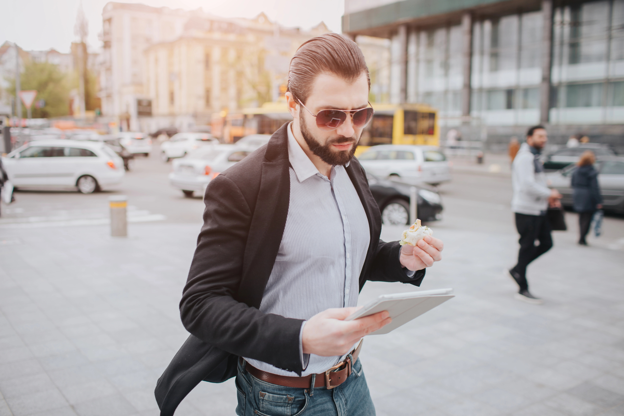 A man with sunglasses and a beard walks on a busy urban street holding a tablet in one hand and eating a doughnut with the other. Cars and people moving in the background under a clear sky with sunlight shining.