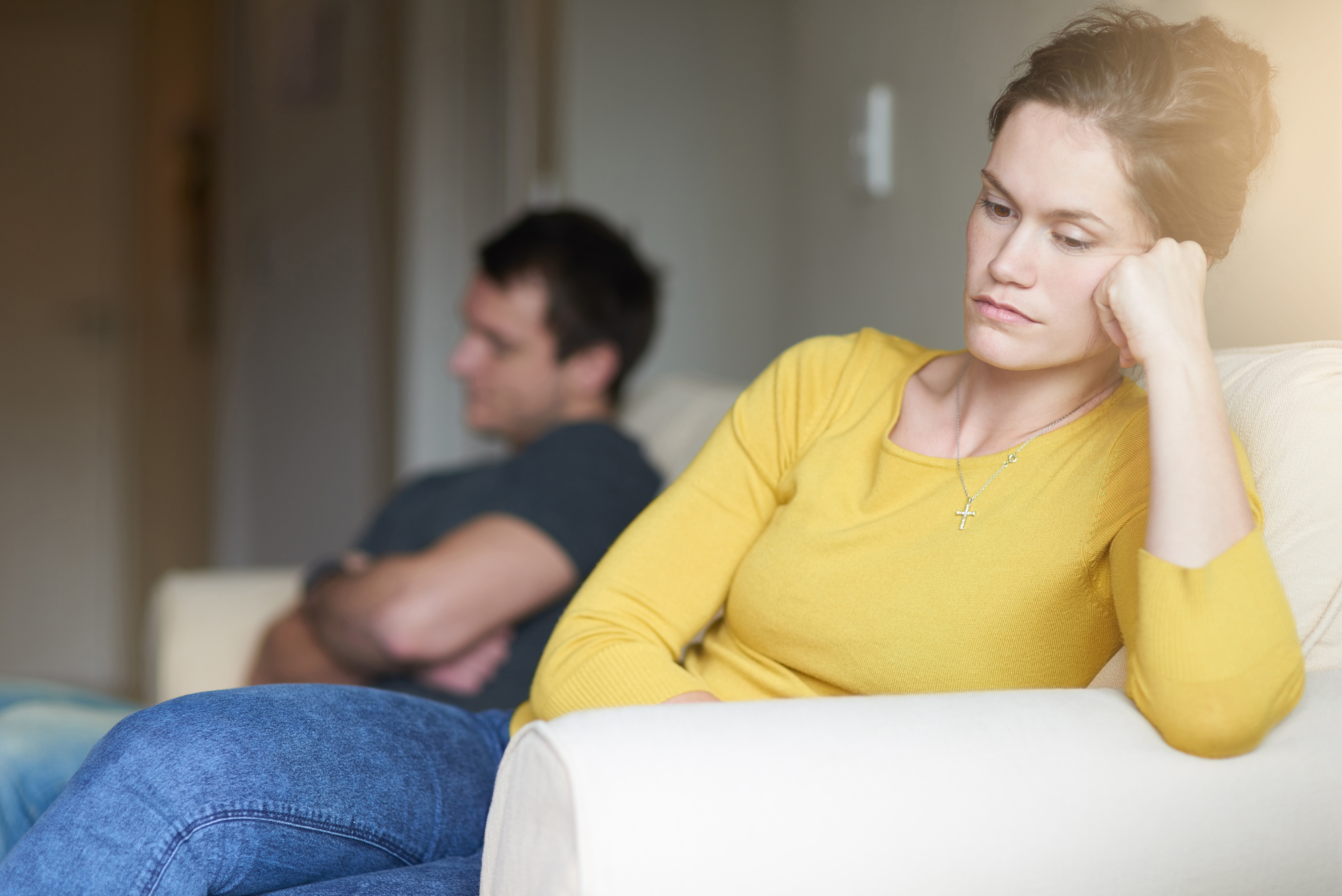 A woman in a yellow sweater and jeans sits on a couch with a thoughtful, concerned expression, resting her chin on her hand. A man in a dark shirt and jeans sits on another couch in the background, his arms crossed, looking away. The setting appears tense.