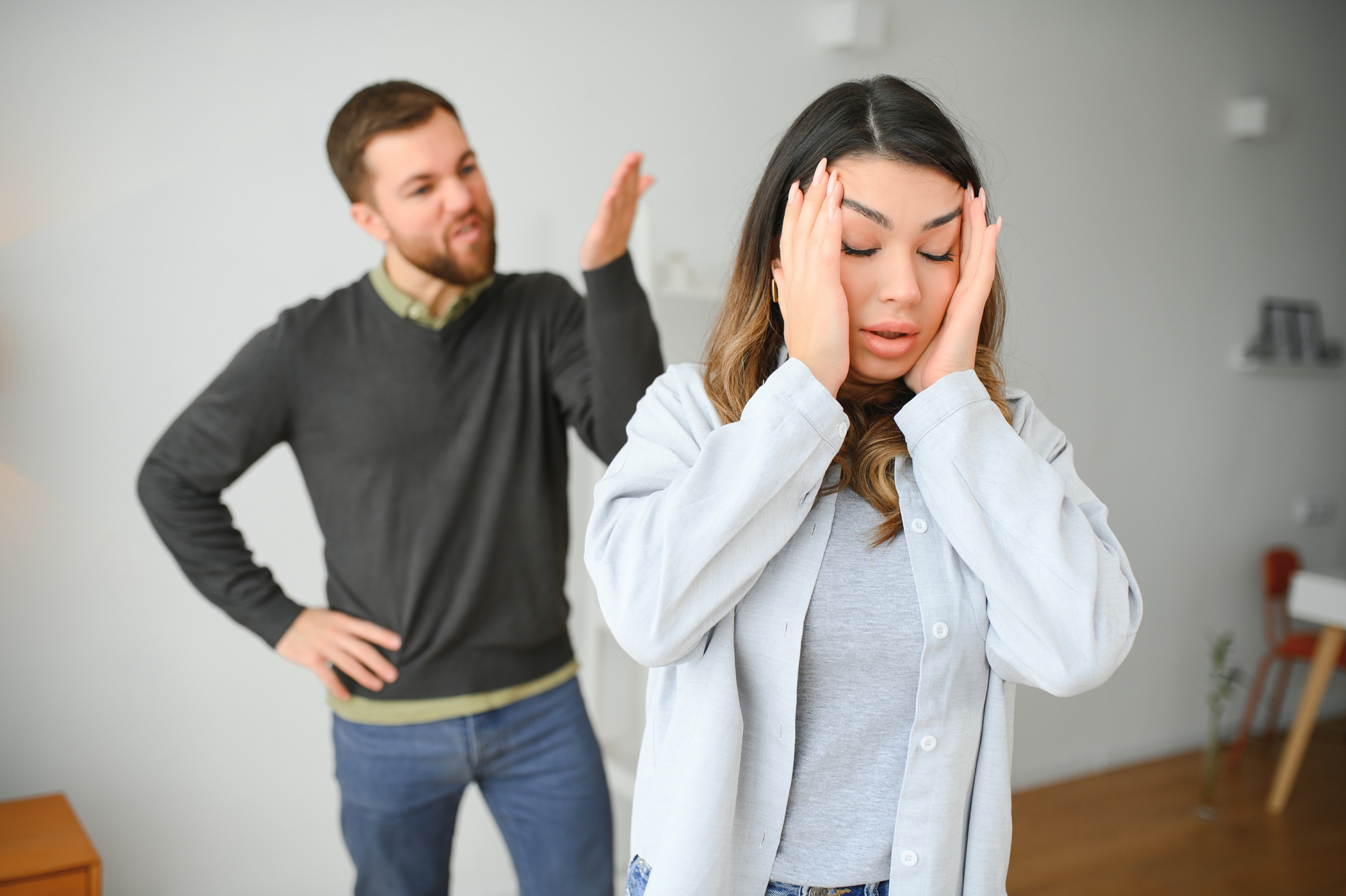 A woman with her hands on her temples appears frustrated while a man behind her raises his hand and seems angry. Both are indoors in a room with white walls and minimal furniture.