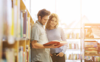 A man and woman stand in a sunlit library, smiling and reading a book together. The man is holding the book open, and both are casually dressed. Shelves filled with books are visible in the background.