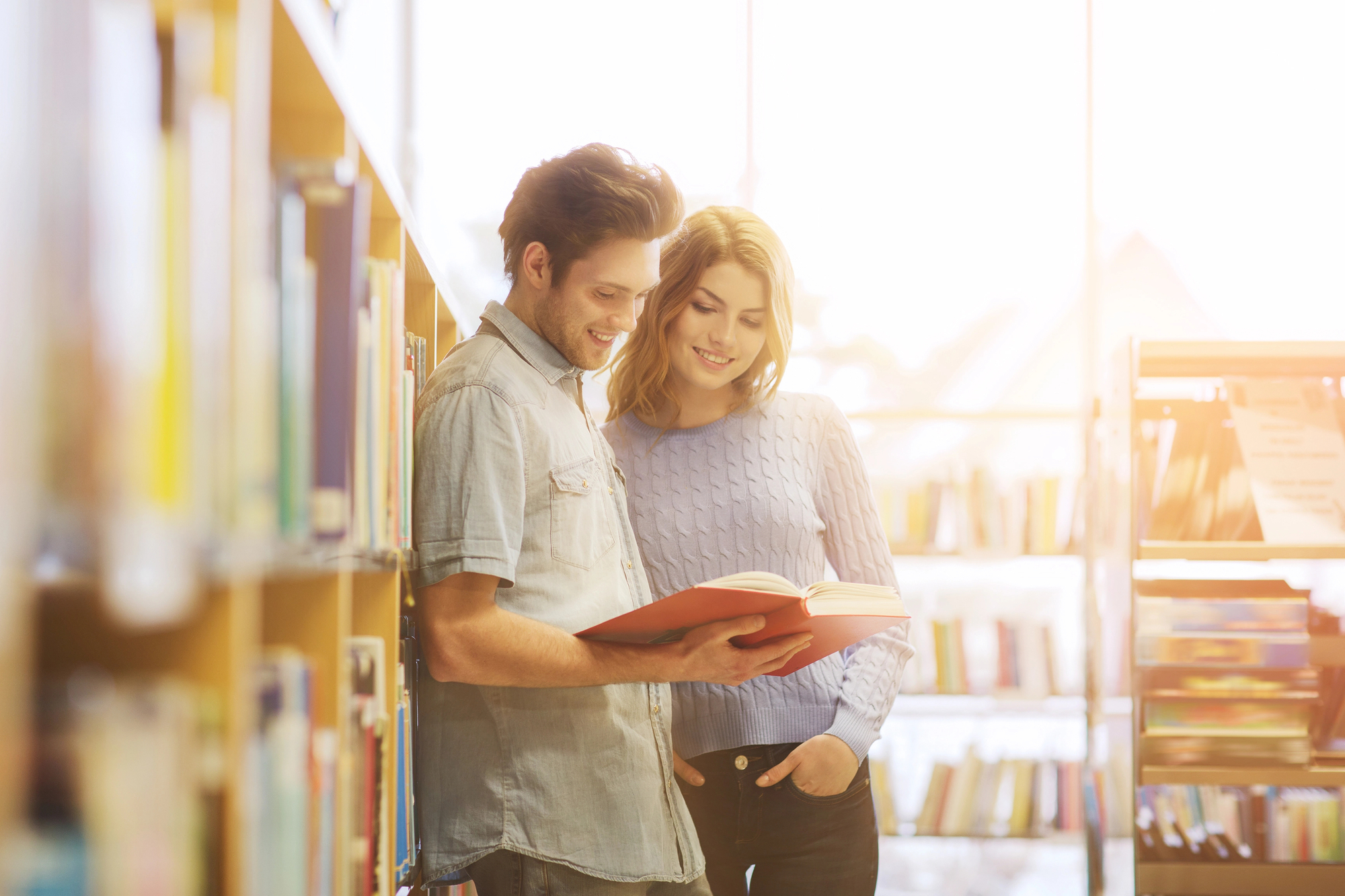 A man and woman stand in a sunlit library, smiling and reading a book together. The man is holding the book open, and both are casually dressed. Shelves filled with books are visible in the background.