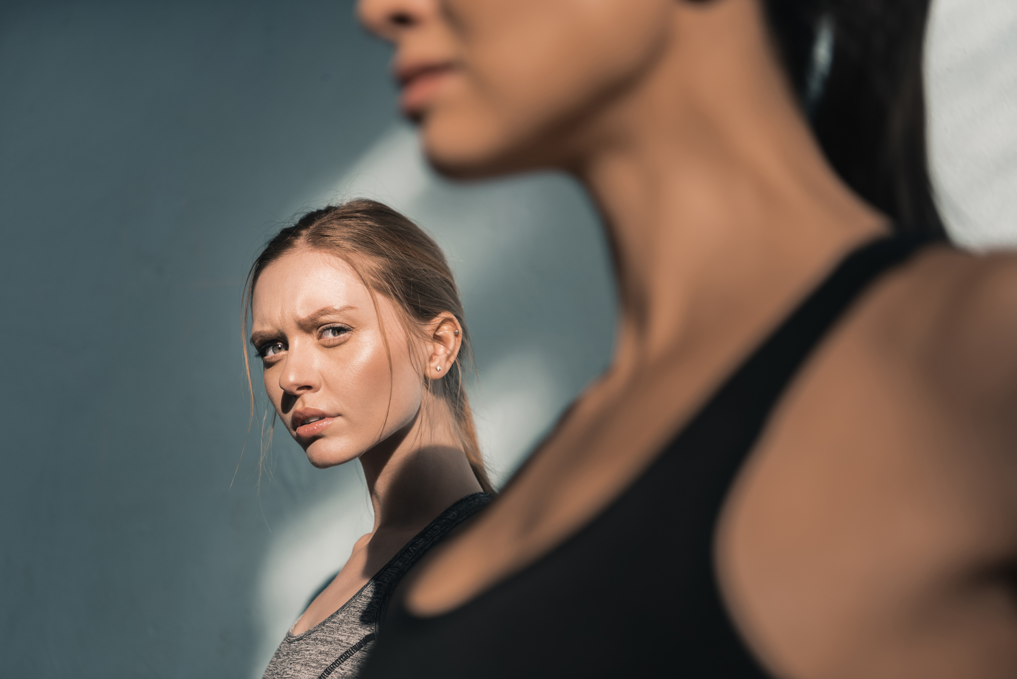 Close-up of two women, one in sharp focus and the other blurred in the foreground. The woman in focus has a serious expression and is wearing a gray top, while the blurred woman in the foreground wears a black top. The lighting creates shadows on their faces.