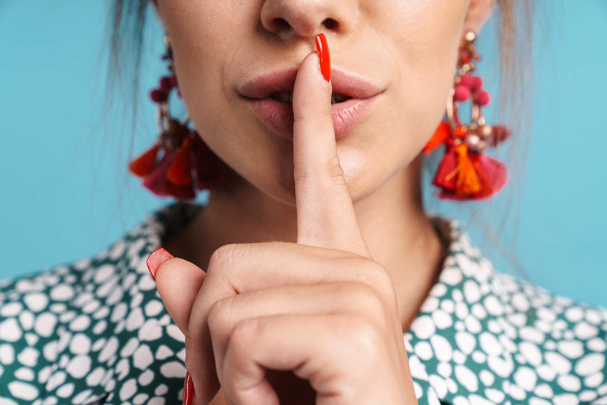 A person with long red nails and wearing colorful earrings is holding their finger to their lips, signaling for silence. They are dressed in a patterned shirt with a blue background.