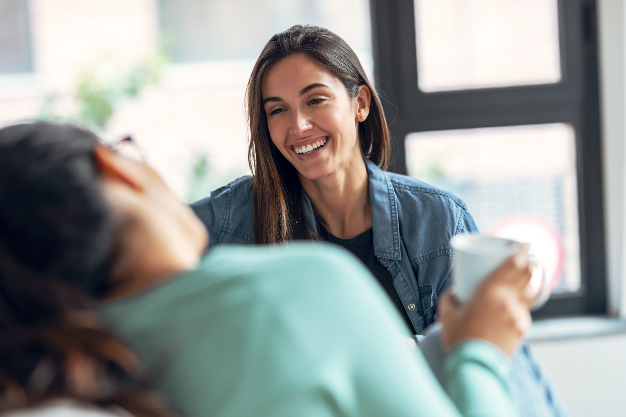 Two people sitting indoors, engaging in a relaxed conversation. One person with long brown hair in a denim shirt is smiling warmly at the other, who is holding a white mug. They are seated near a large window with blurred greenery outside.