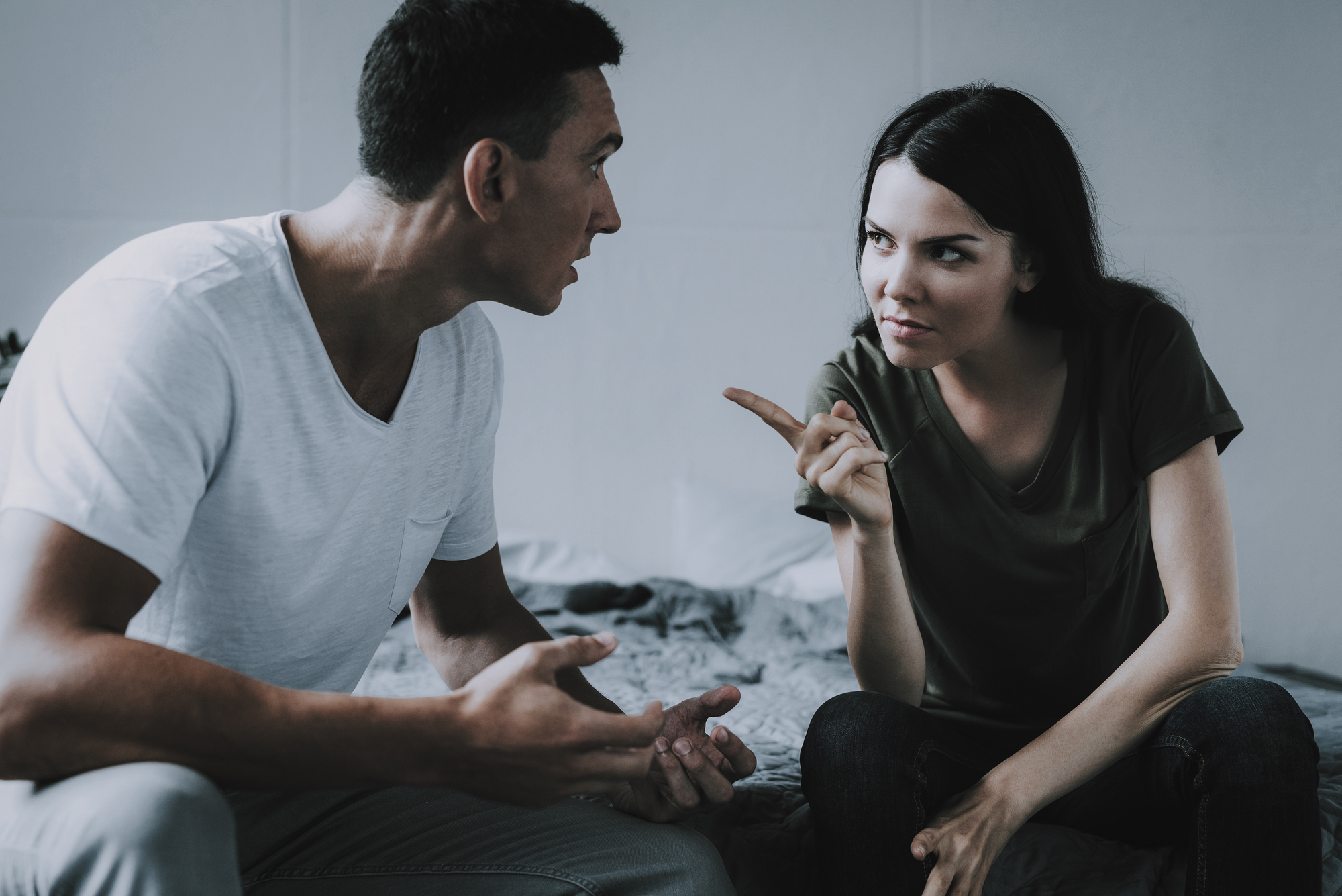 A man and a woman are sitting on a bed facing each other, engaged in an intense conversation. The woman points her finger at the man in a stern manner, while the man gestures with both hands. Both look serious and focused. The background is a simple, light-colored room.