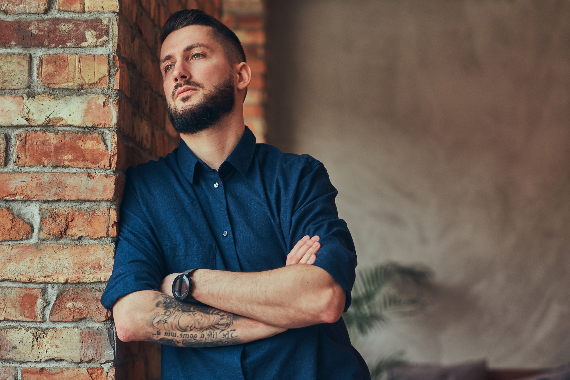 A man with a beard and a tattoo on his forearm is leaning against an exposed brick wall with his arms crossed. He is wearing a dark blue shirt and looking off into the distance with a contemplative expression. The background is blurred, featuring some greenery.