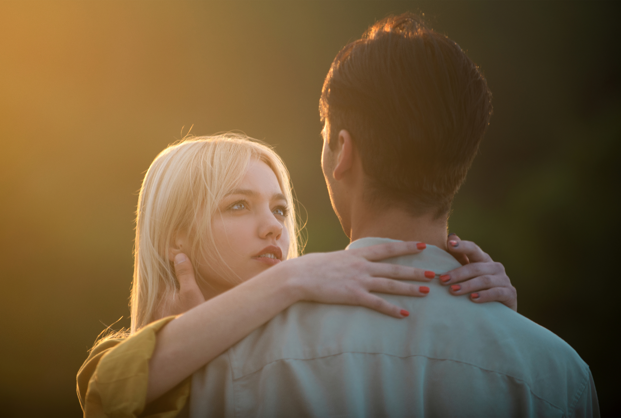 A blonde woman with red nail polish embraces a man with dark hair, gazing into his eyes. They are illuminated by a warm, golden light, likely from a sunset, creating a romantic and intimate atmosphere. The background is blurred and dark.