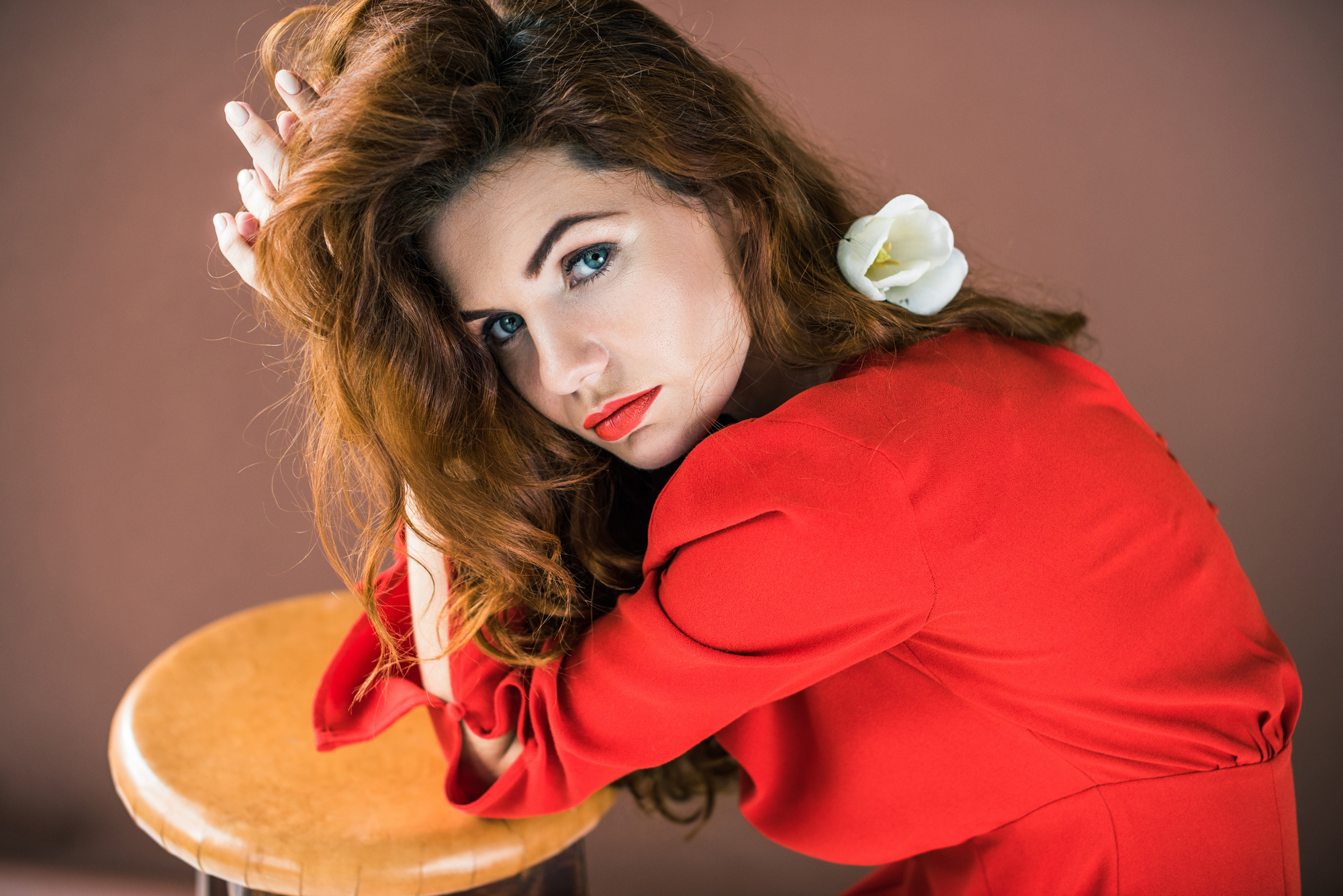A woman with long red hair and a white flower tucked behind her ear is leaning on a wooden stool. She is wearing a red dress and gazing intently at the camera, her hands resting on top of the stool. The background is a warm, muted color.