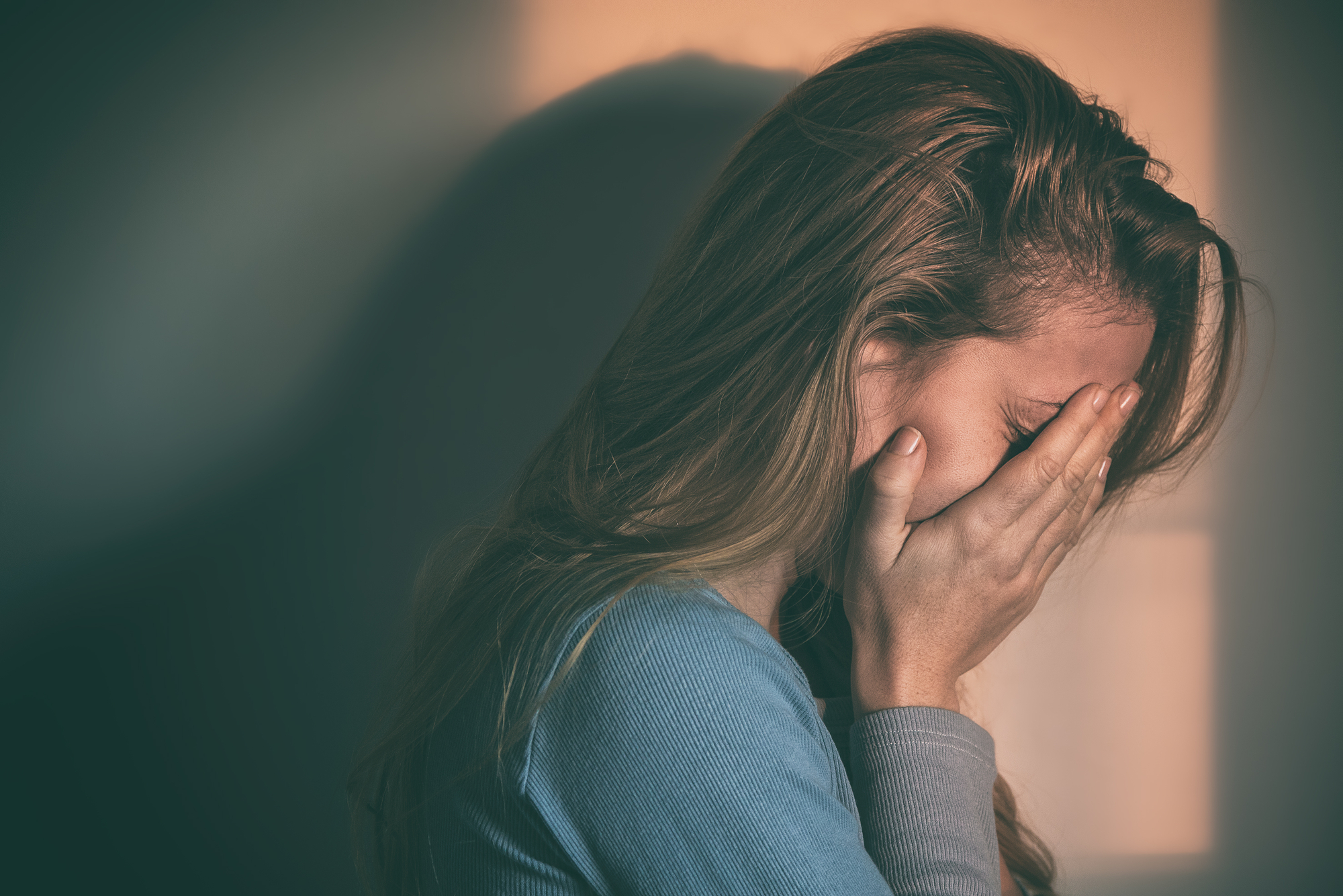 A woman with long hair, wearing a blue shirt, covers her face with her hands in an expression of distress. She appears to be emotional or upset, and the background is softly lit with a warm light.
