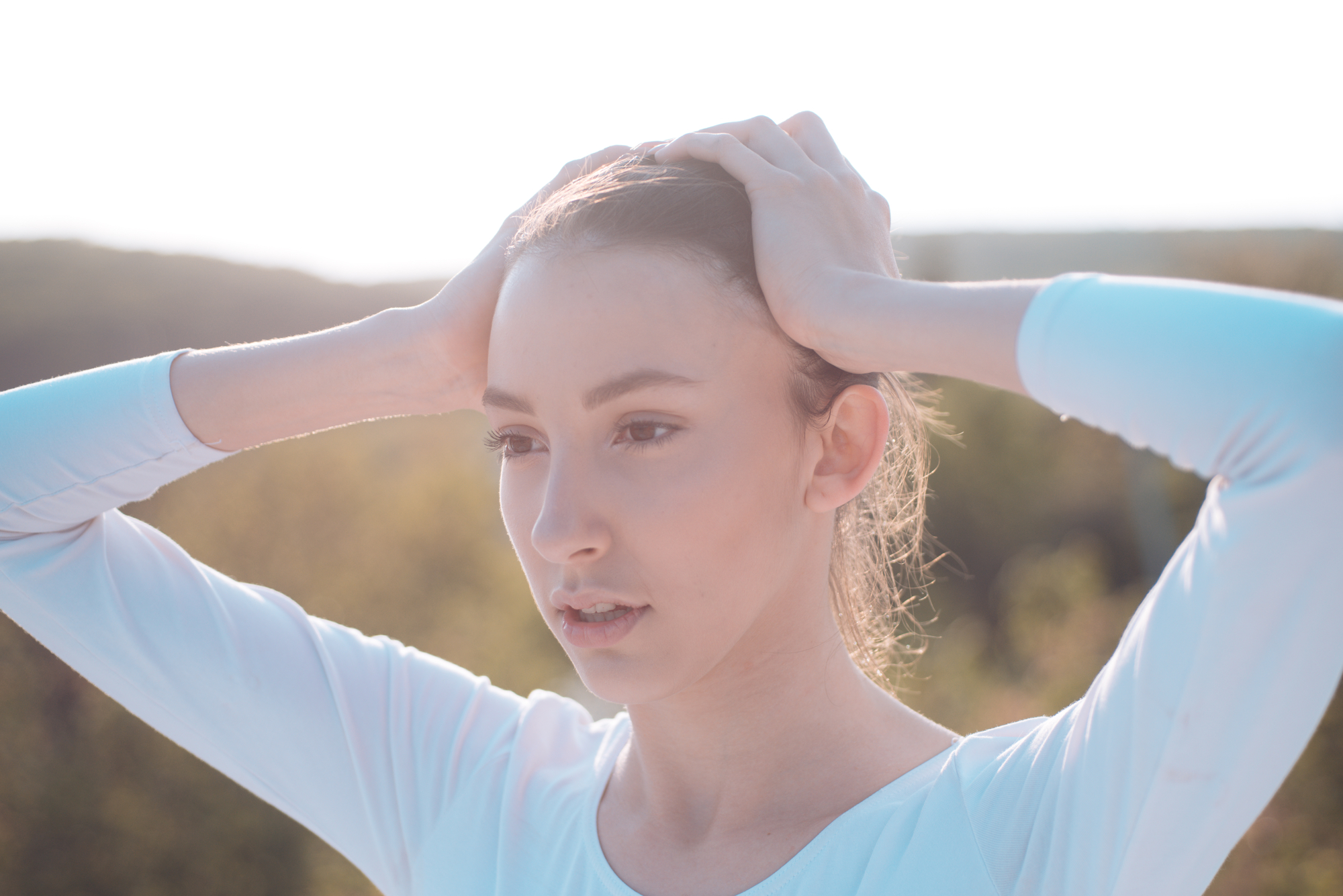 A young person with long hair pulled back is outdoors, wearing a light-colored long sleeve top. They have their hands placed on their head and appear to be looking into the distance. The background is softly blurred, indicating a natural setting.