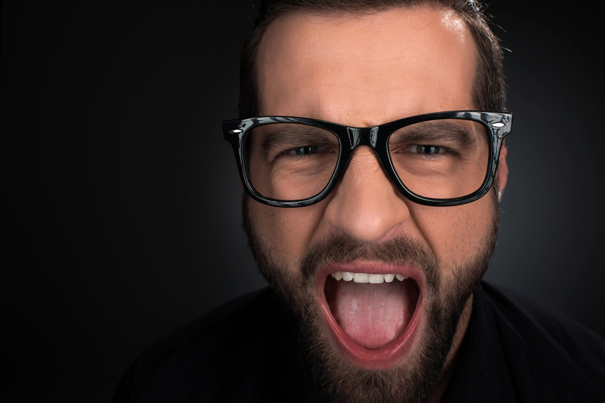 A close-up shot of a man with a beard and glasses, his mouth wide open as if shouting or expressing strong emotion. The background is dark, emphasizing his facial expression and the details of his face.