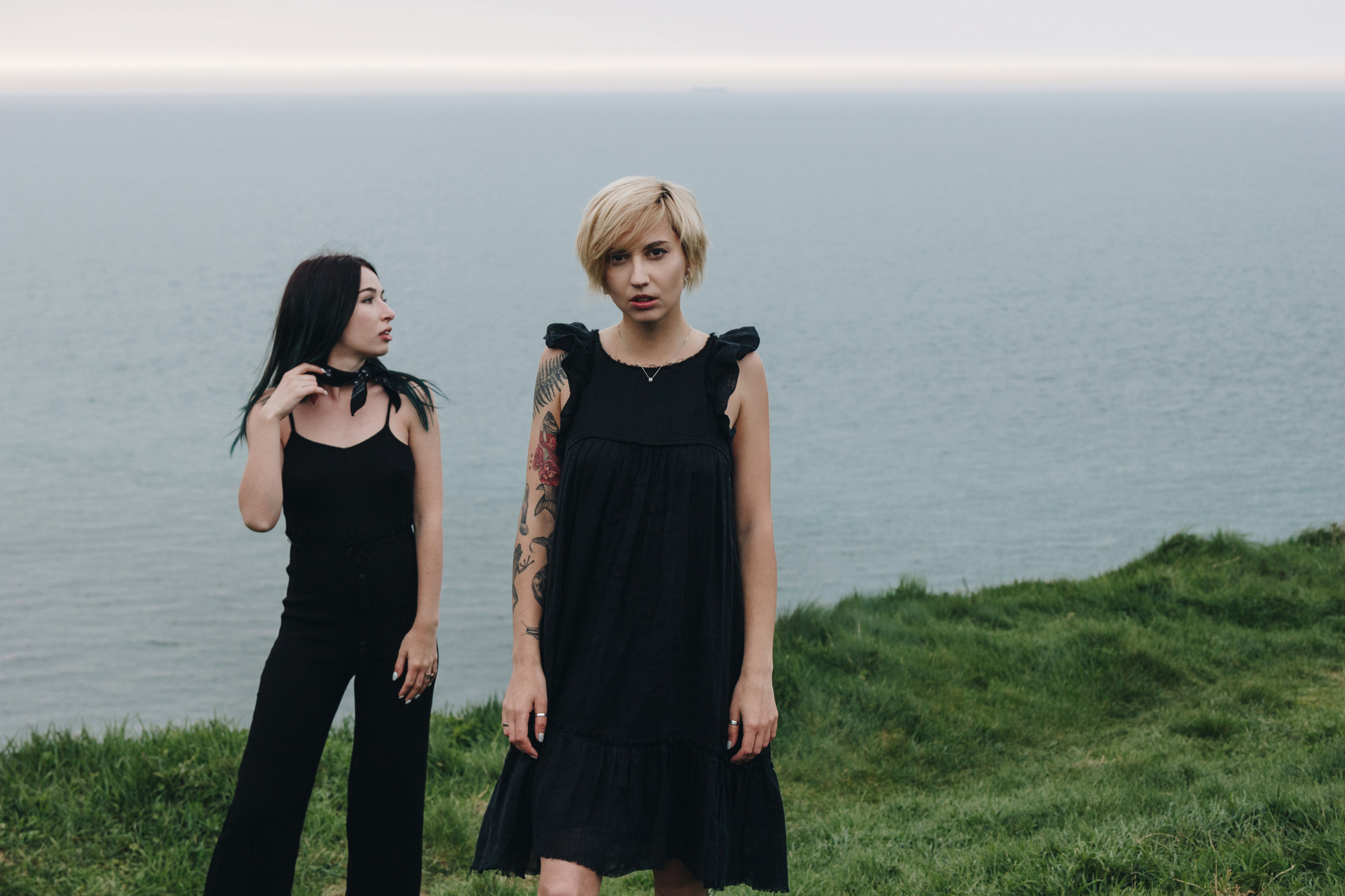 Two women stand on a grassy cliff overlooking a calm sea. The woman on the left, with long black hair, wears a black sleeveless top and pants, looking to her right. The woman on the right, with short blonde hair and a tattooed arm, wears a black dress, facing the camera.