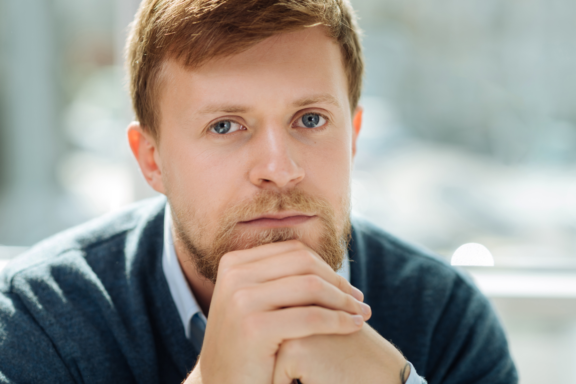 Young man with short, light brown hair and a beard looking directly into the camera. He is resting his chin on his hands and wearing a blue sweater over a collared shirt. The background appears to be out of focus, suggesting a bright and airy indoor setting.