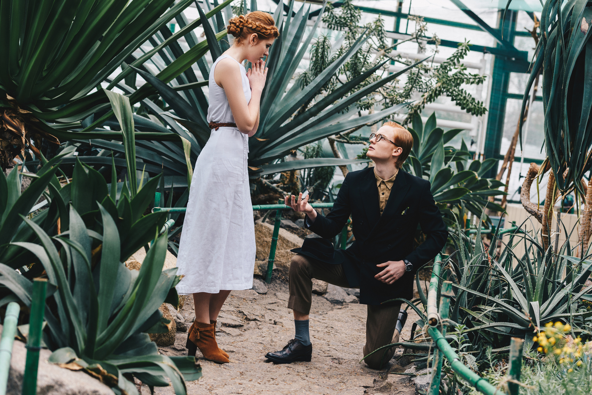A man in a suit kneels and proposes to a surprised woman in a white dress and brown boots. They are surrounded by lush green plants inside a greenhouse. The woman appears emotional, with her hands clasped near her face.