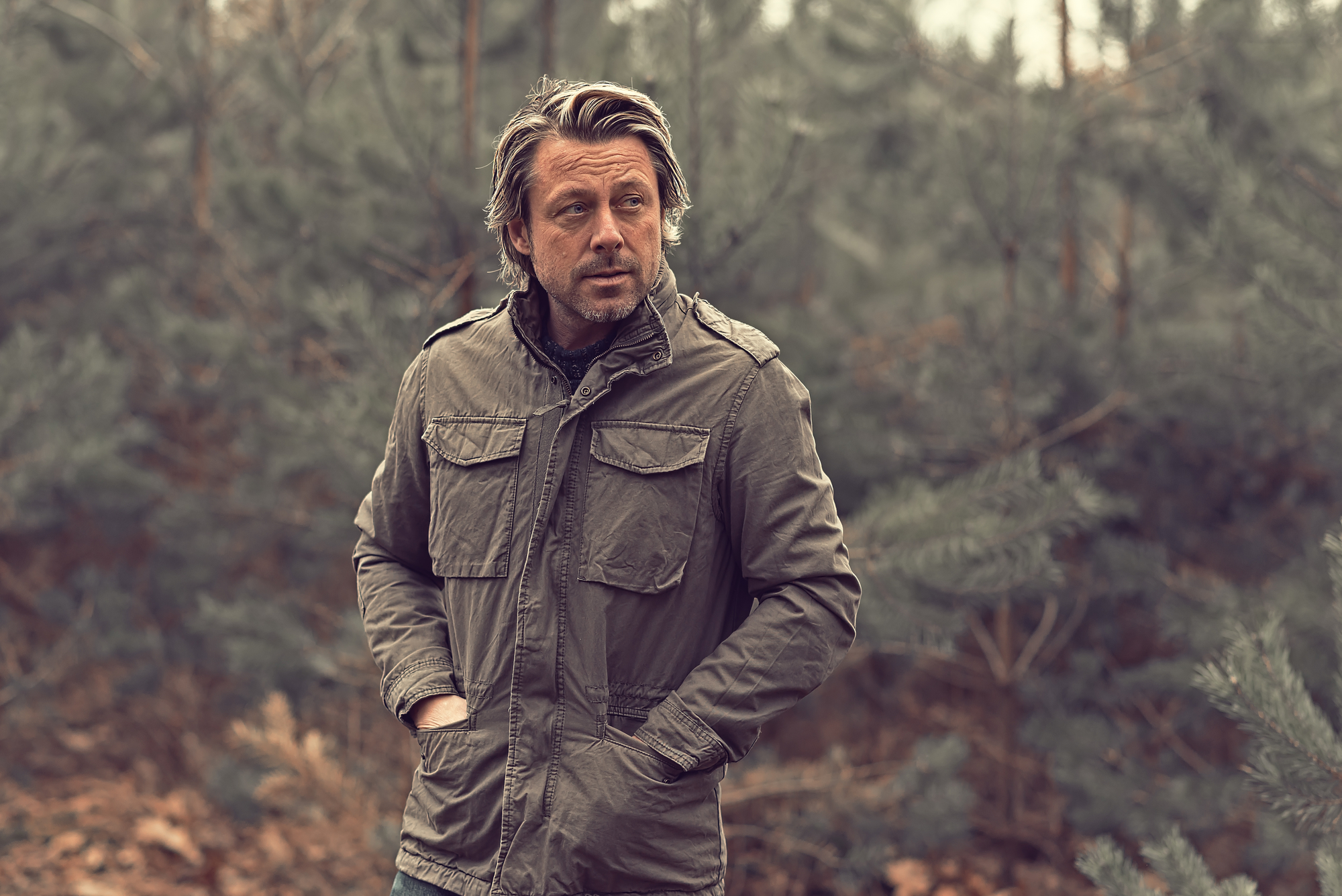A man with light brown hair, wearing a green jacket with his hands in the pockets, stands outdoors among evergreen trees. The background is blurry, emphasizing the forest setting. The man gazes off to the side with a contemplative expression.