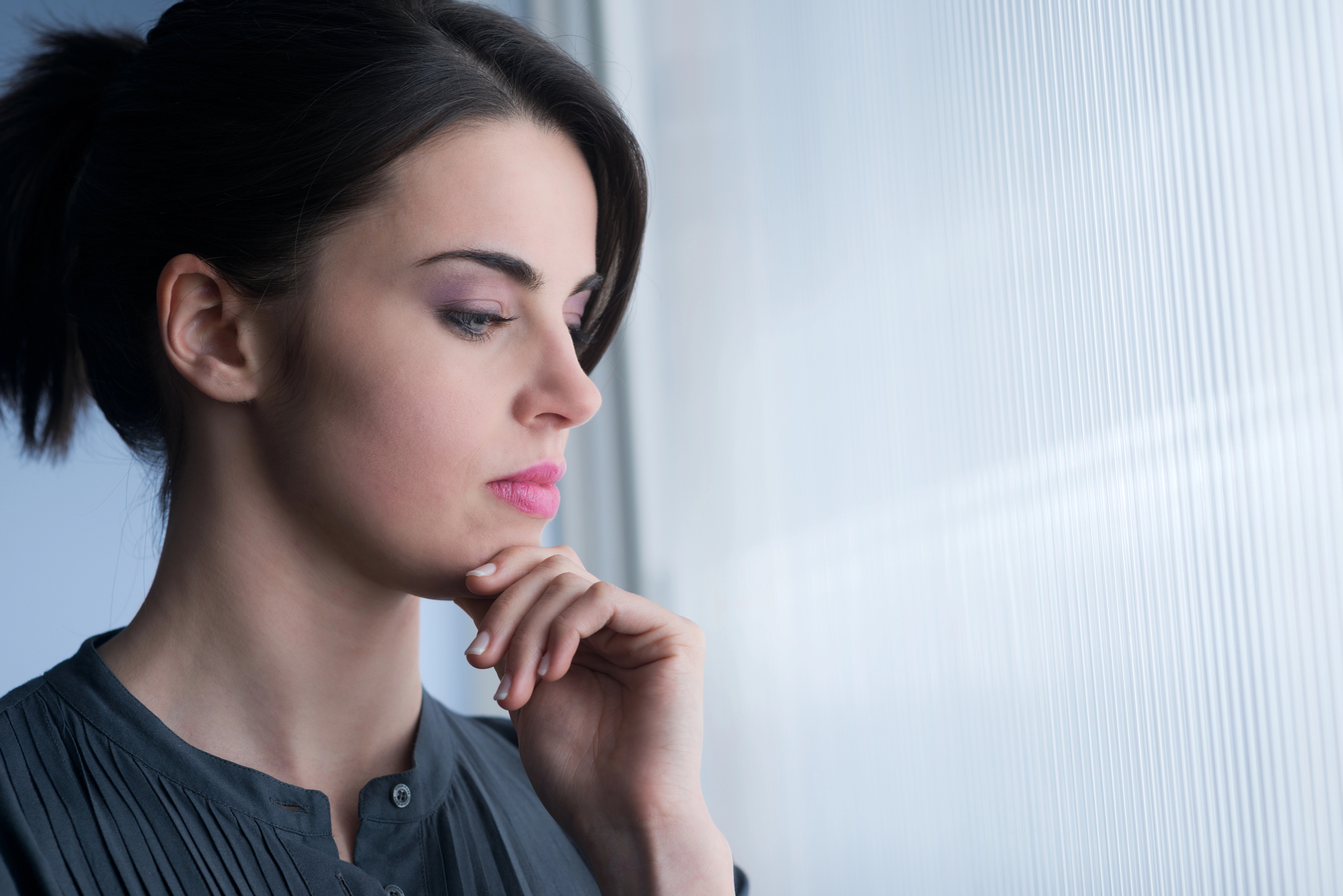 A woman with dark hair pulled back in a ponytail stands by a frosted window, looking out with a pensive expression. She rests her chin lightly on her hand, wearing a dark, long-sleeve top. The light from the window softly illuminates her face.