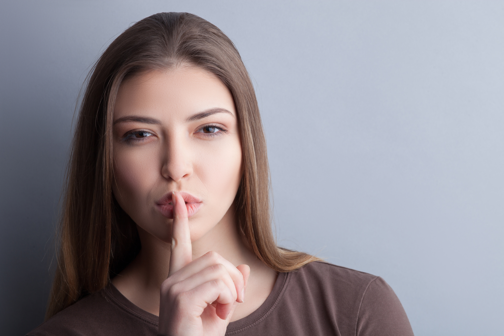 A woman with long brown hair wearing a brown shirt holds her index finger to her lips in a 'shushing' gesture, suggesting silence. She has a calm expression and is standing against a plain gray background.