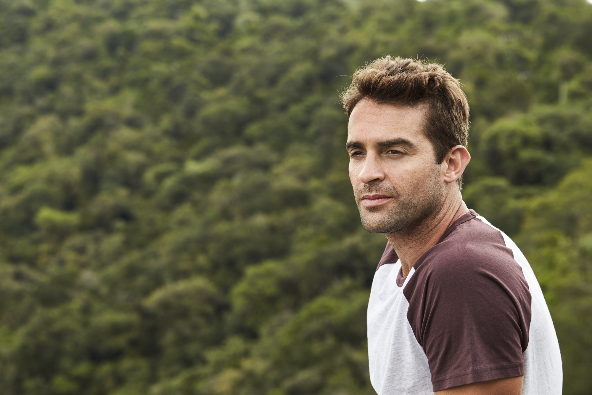 A man with short brown hair, wearing a white and maroon shirt, gazes thoughtfully to the side. He is outdoors, with a lush green forest visible in the background.