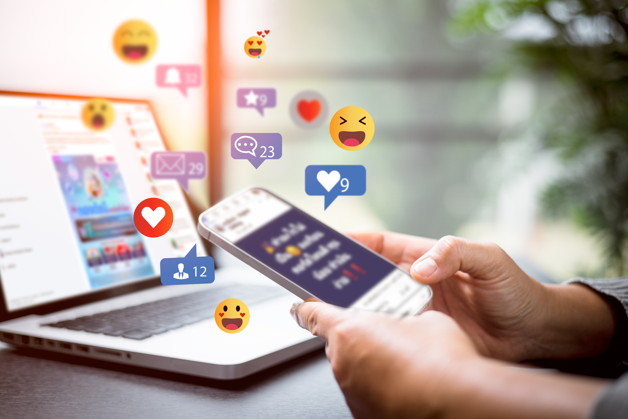 A person holds a smartphone displaying social media notifications of likes, comments, and emojis. A laptop with an open social media page is in the background, also showing notifications. The scene is brightly lit with a blurred background of greenery.