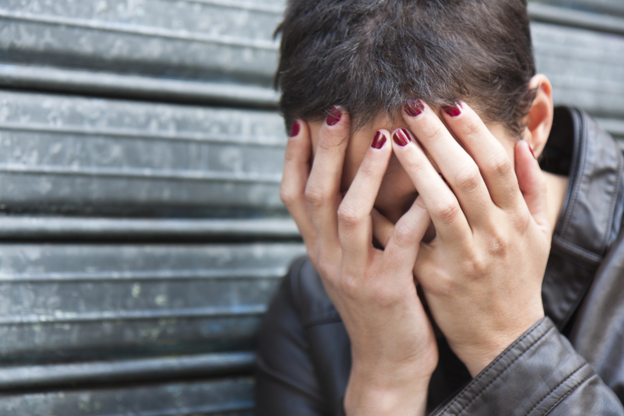 A person with short hair and red-painted nails rests their face in their hands, appearing distressed. They are wearing a dark leather jacket and are positioned in front of a corrugated metal wall.