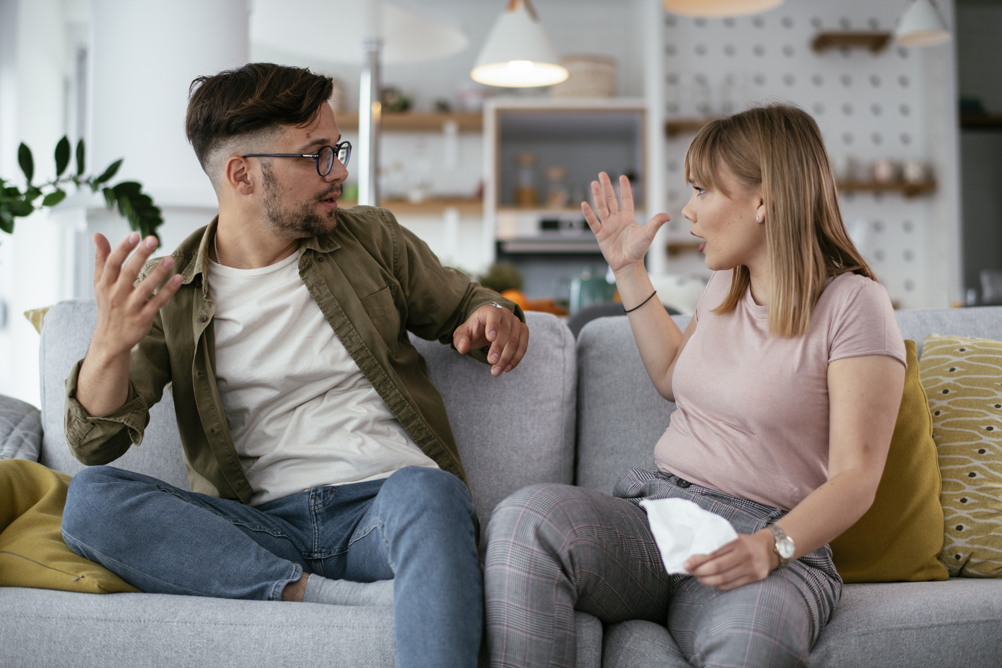A man and woman are sitting on a gray couch in a modern living room, having an animated conversation. Both have raised hands and expressive gestures, indicating a heated discussion. The woman holds a tissue, and both appear emotionally engaged.