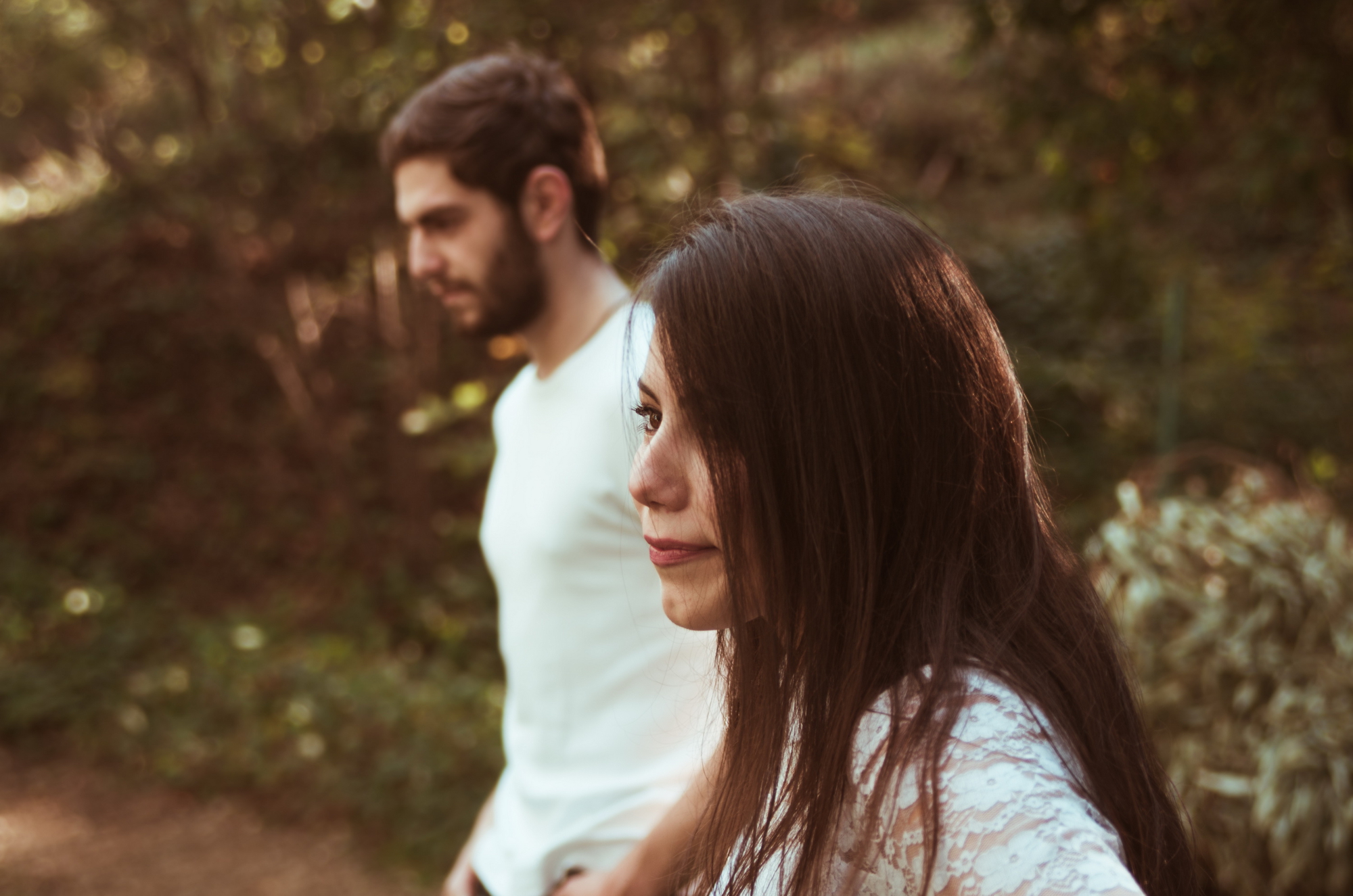 A man and woman walk outdoors with foliage in the background. The man, in a white shirt, has dark hair pulled back and a beard. The woman, in a white lace top, has long dark hair. The focus is on the woman’s profile, with the man slightly blurred in the background.