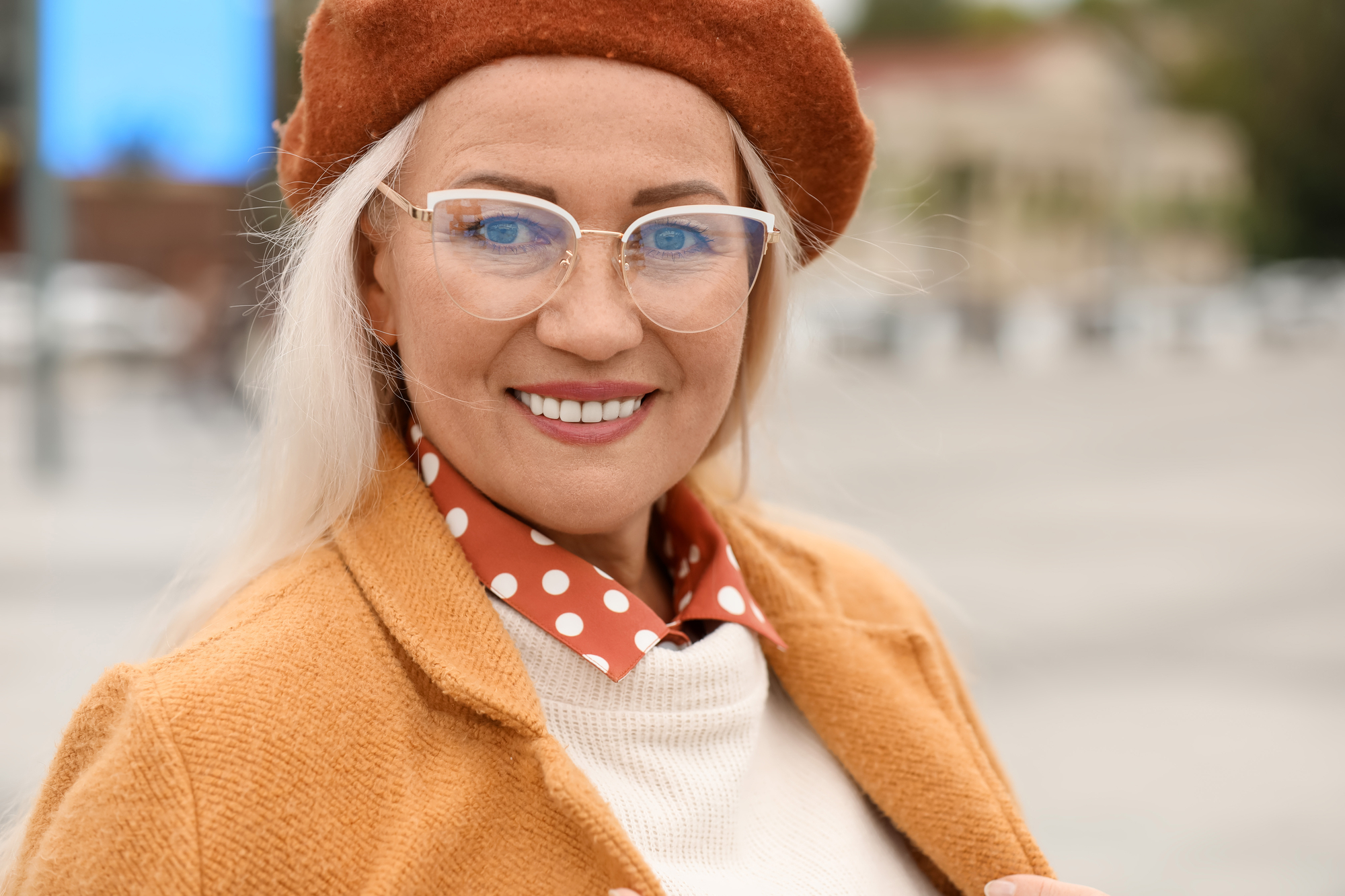 A woman with long blonde hair smiles at the camera. She is wearing a brown beret, clear glasses, and a mustard-colored coat over a white sweater and red polka-dotted shirt. The background is an outdoor scene with blurred buildings and trees.