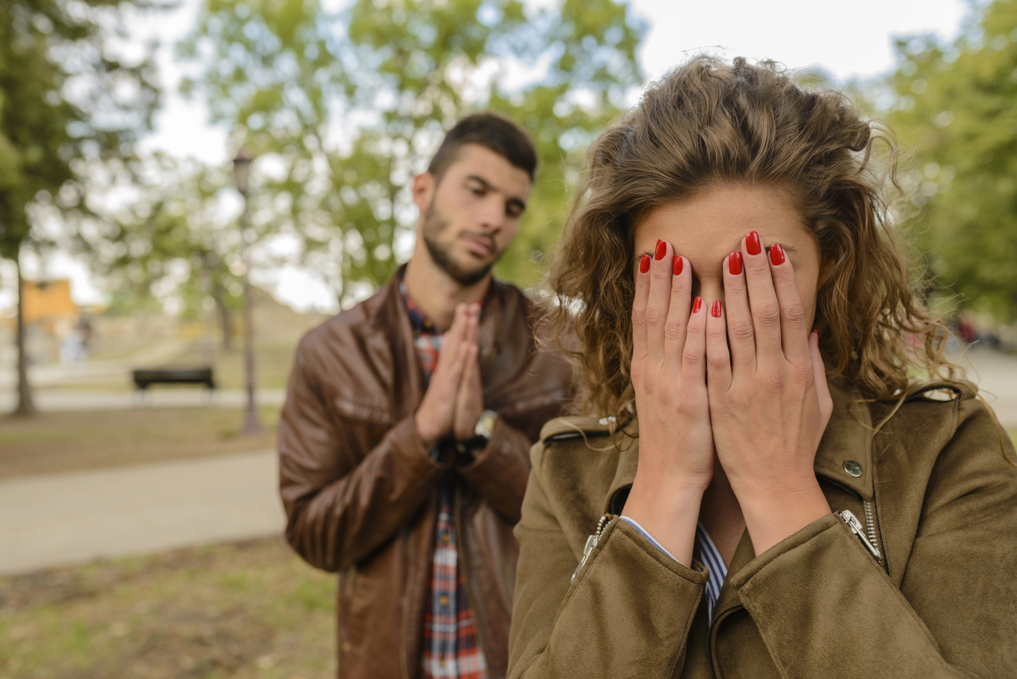 A woman with curly hair wearing a jacket covers her face with her hands while standing outdoors. Behind her, a man with short hair wearing a brown leather jacket and plaid shirt clasps his hands together as if pleading or apologizing.