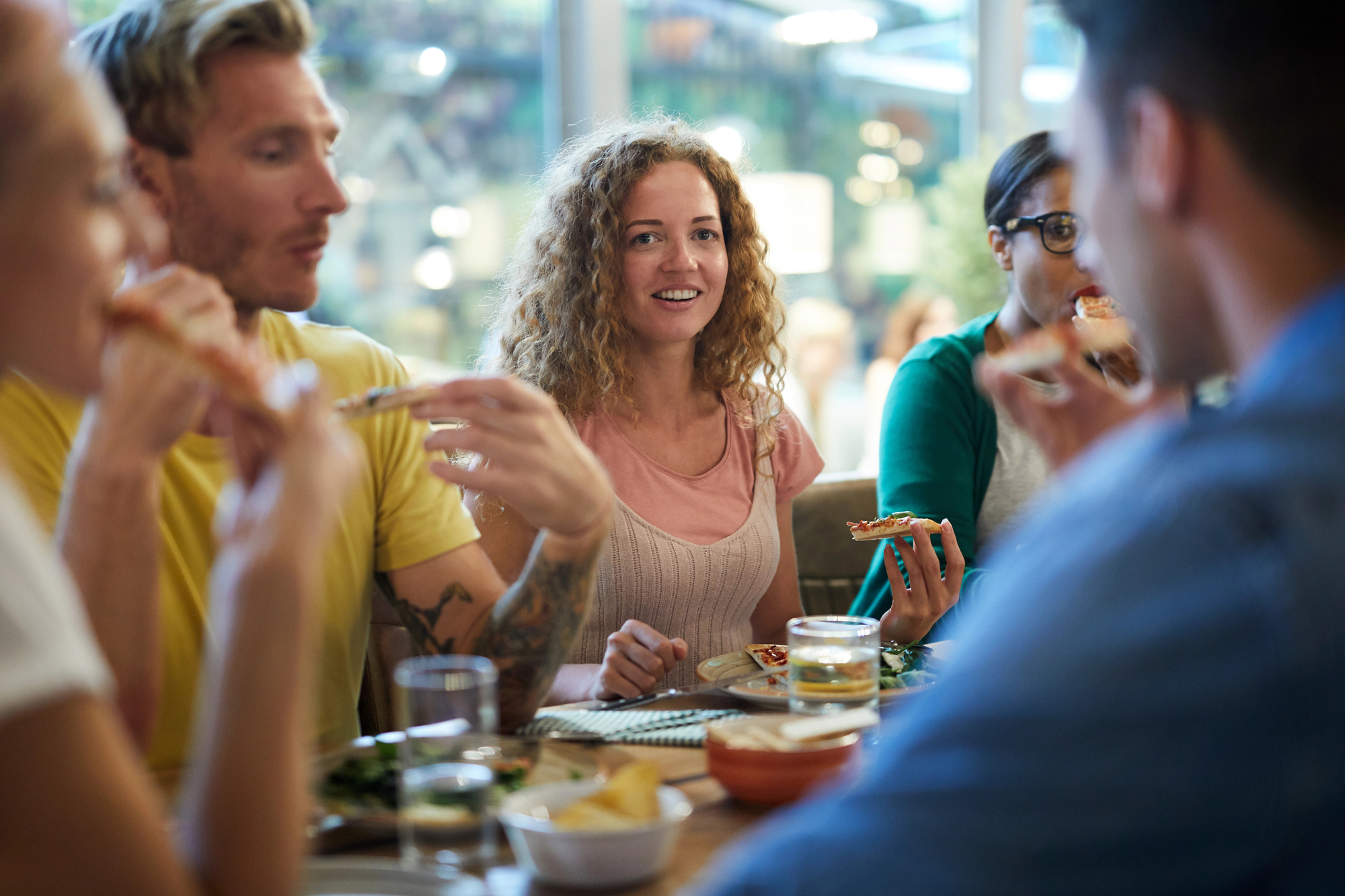 A group of people sitting around a table in a restaurant, eating and talking. A woman with curly hair wearing a pink top and beige vest is smiling in the center, while others around her are holding pieces of pizza. The atmosphere appears lively and social.