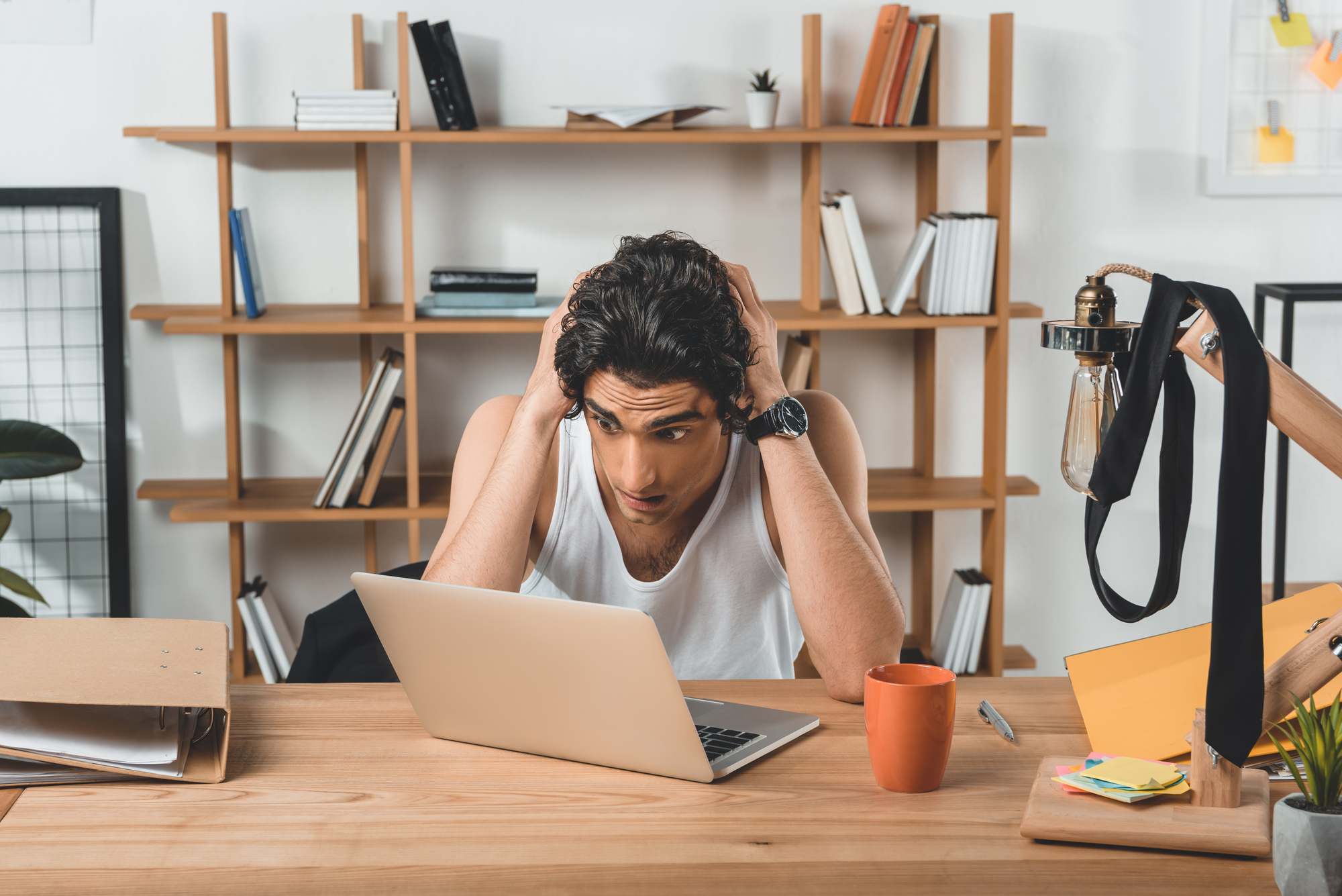 A person with dark hair wearing a white tank top sits at a desk, looking stressed with their hands on their head. In front of them is an open laptop and a mug. The desk is cluttered with various items, and a wooden shelf with books and decor is in the background.