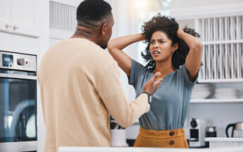 A man and woman are having an intense conversation in a bright kitchen. The woman, with a concerned expression, has her hands on her head. The man speaks to her gesturing with his hand. Various kitchen appliances and items are visible in the background.