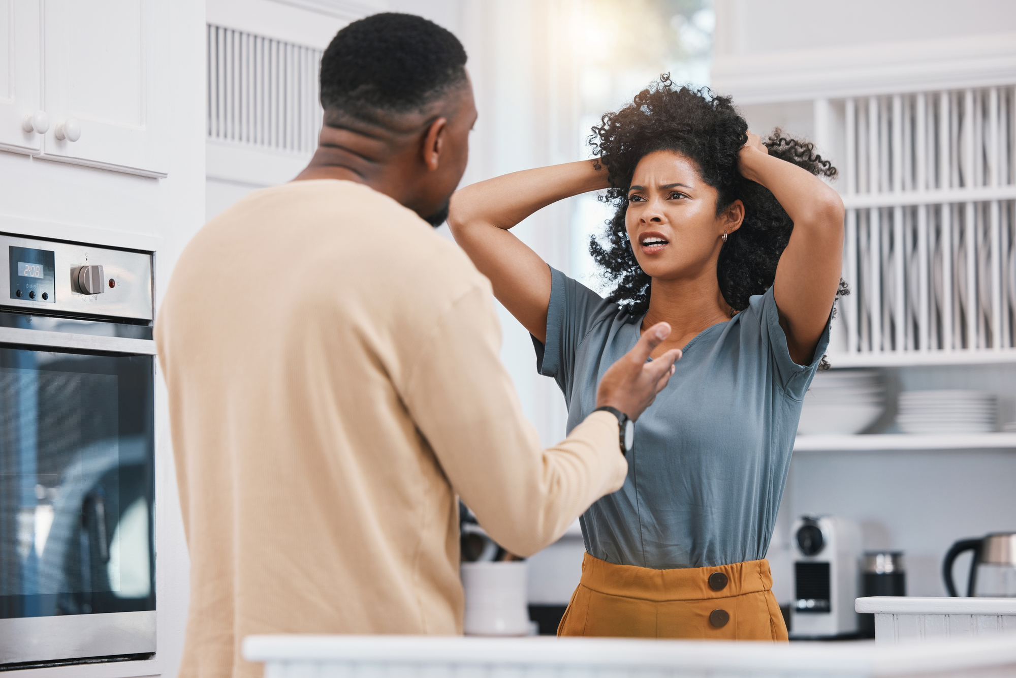 A man and woman are having an intense conversation in a bright kitchen. The woman, with a concerned expression, has her hands on her head. The man speaks to her gesturing with his hand. Various kitchen appliances and items are visible in the background.