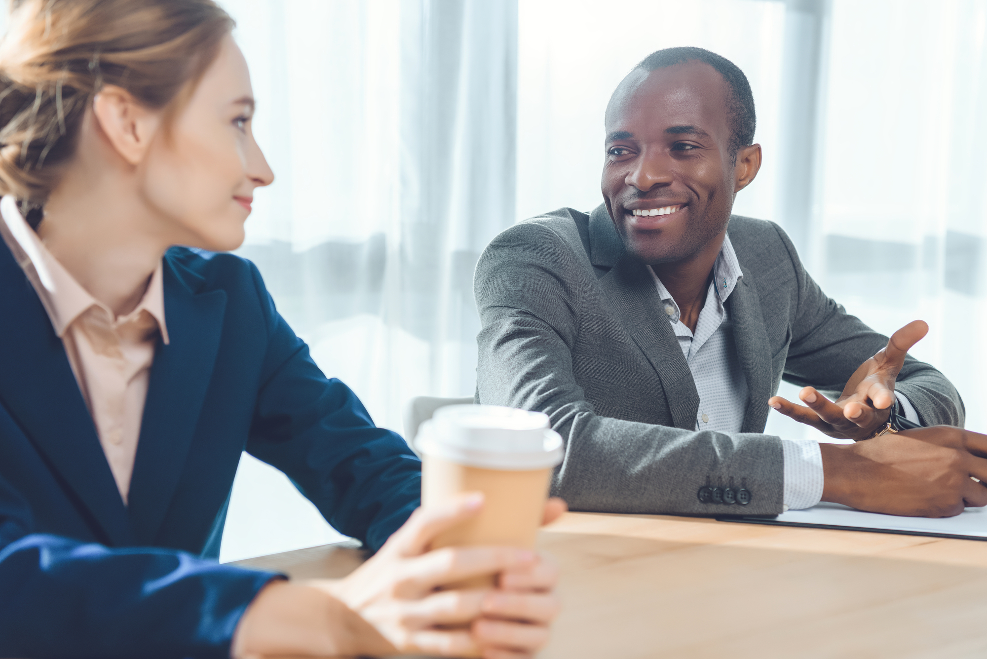 Two colleagues having a friendly conversation at a table. The woman is holding a coffee cup while smiling, and the man is engaged in the discussion, gesturing with one hand. Both are dressed in business attire, with bright natural light coming through the windows.