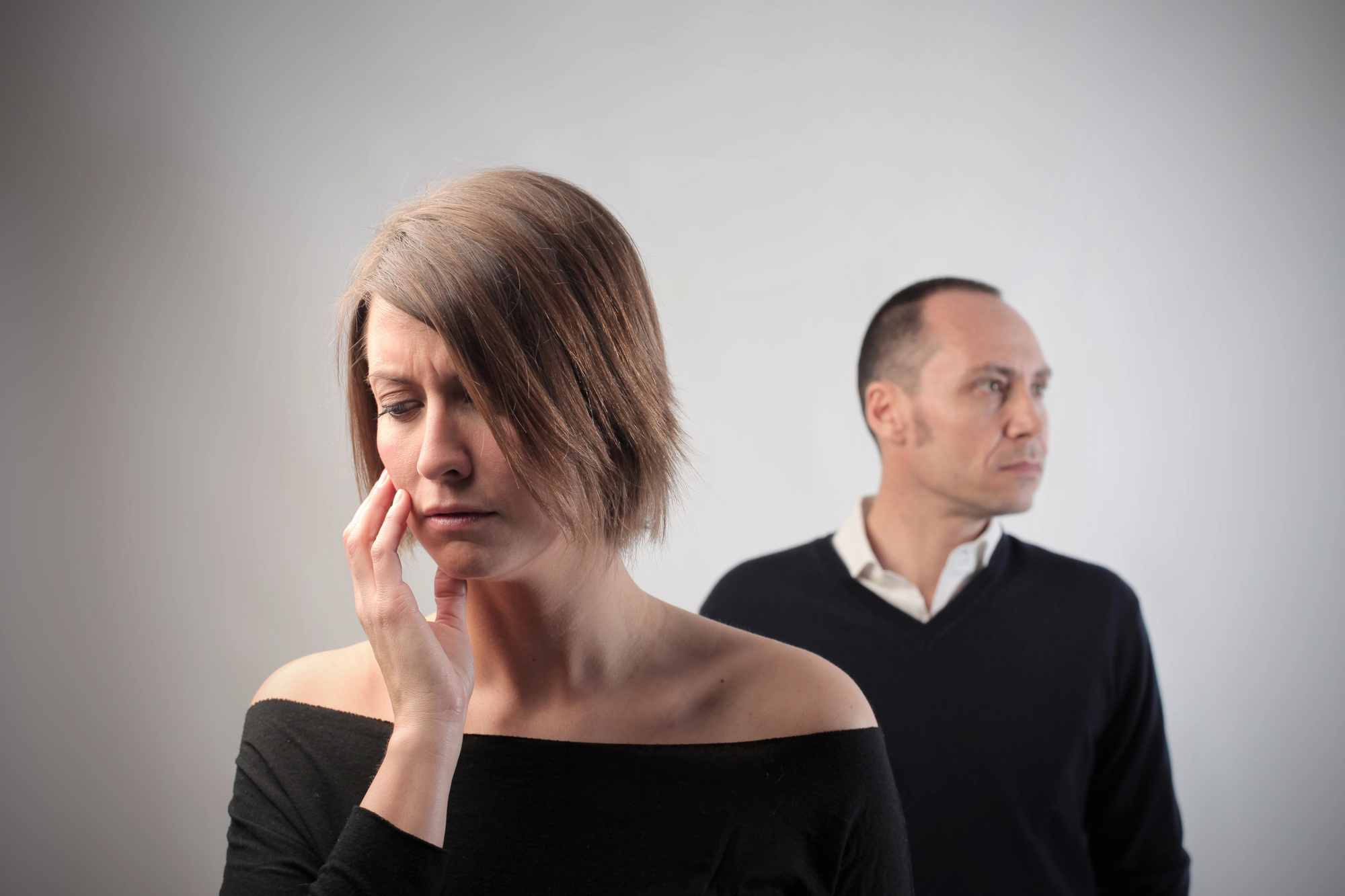 A woman with short, brown hair appears upset, touching her cheek with her hand and looking down. In the background, a man with short hair, wearing a dark sweater and white shirt, stands slightly out of focus, facing away with a stern expression.
