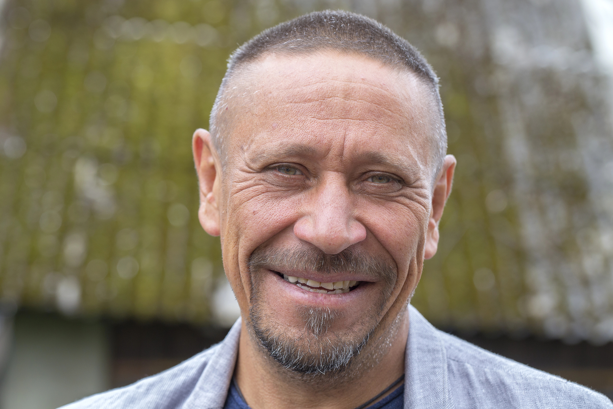 A middle-aged man with a short buzz cut hairstyle smiles warmly at the camera. He has a neatly trimmed goatee and is wearing a casual outfit with a light jacket. The background is slightly blurred, showing what appears to be a moss-covered roof.