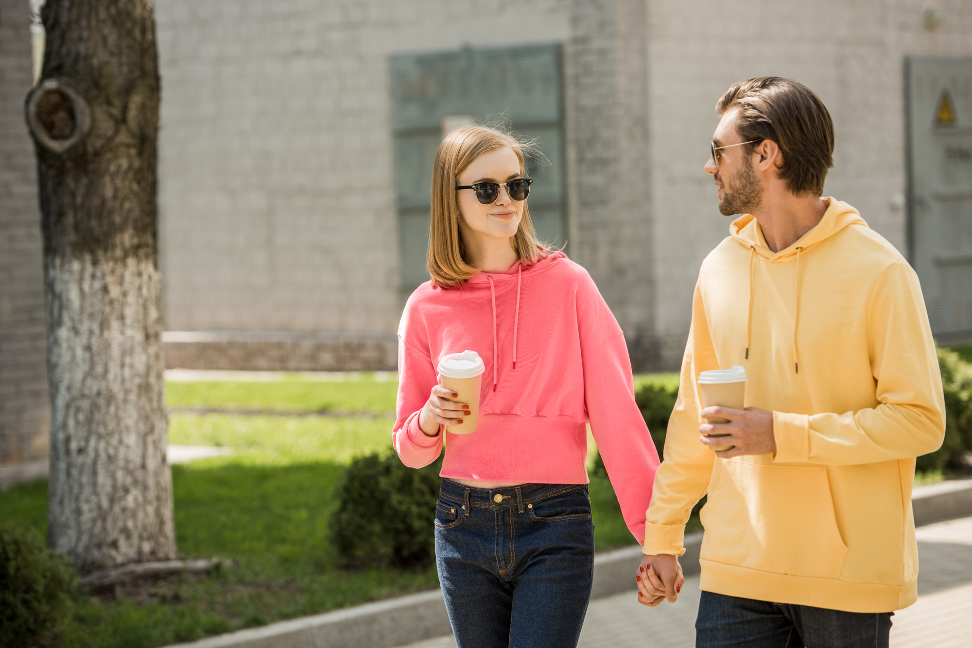 A young couple holding hands while walking outdoors and drinking coffee. Both are wearing sunglasses, with the woman in a pink hoodie and jeans, and the man in a yellow hoodie and jeans. They appear to be enjoying a pleasant day in a park-like setting.