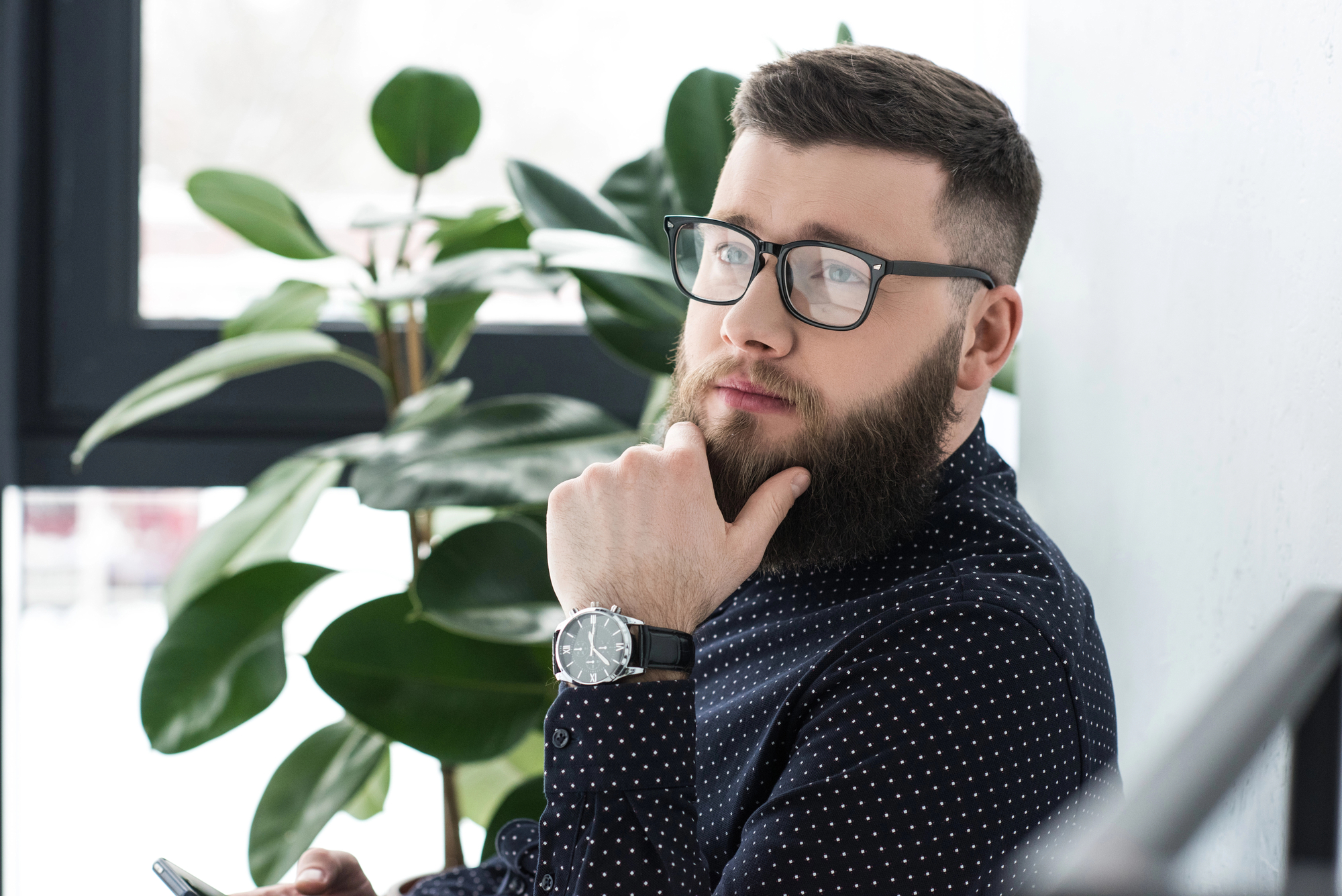 A bearded man with glasses, wearing a dark polka-dot shirt and a wristwatch, is sitting near a window with a potted plant behind him. He is looking thoughtfully into the distance, with one hand resting on his chin.