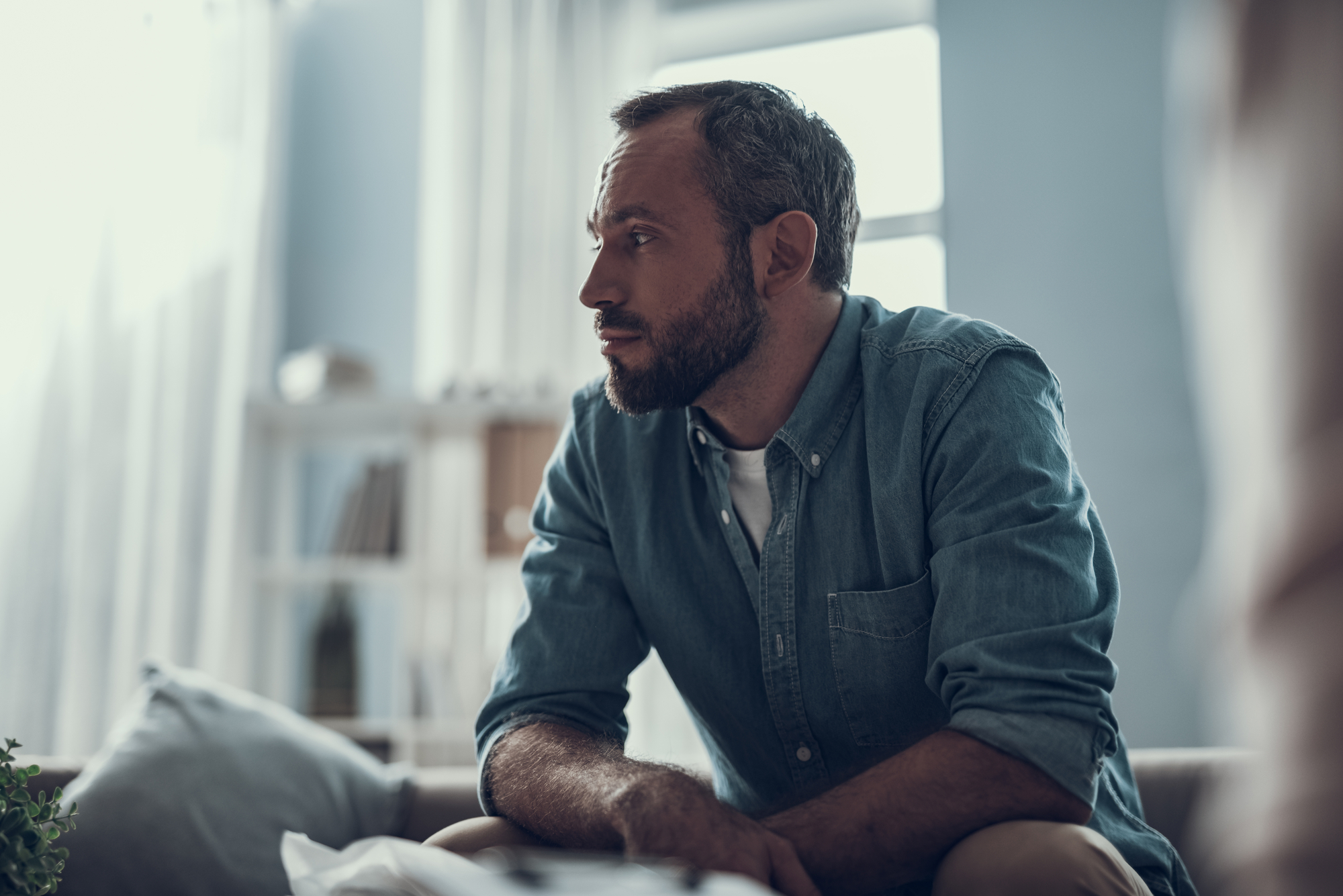 A man with short hair and a beard, wearing a denim shirt, is sitting on a couch in a brightly lit room. He is looking pensively to the side. The background features shelves with books and decor items.