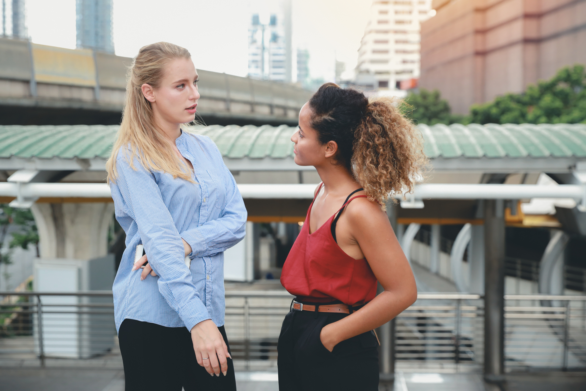 Two women are having a conversation outdoors near an urban overpass. One woman, with blonde hair, is wearing a blue long-sleeve shirt and has her arms crossed. The other woman, with curly hair, is wearing a red sleeveless top and dark pants, and is gesturing with her hands.