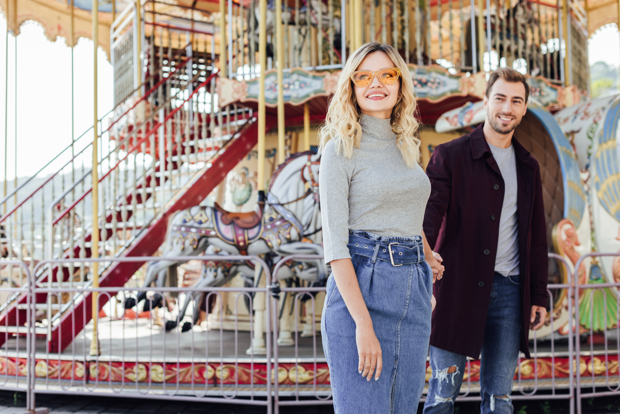A smiling woman wearing sunglasses and a gray turtleneck stands in front of a colorful carousel, holding hands with a man in a dark coat. The man has light brown hair and a beard. The carousel features ornate decorations and horses.